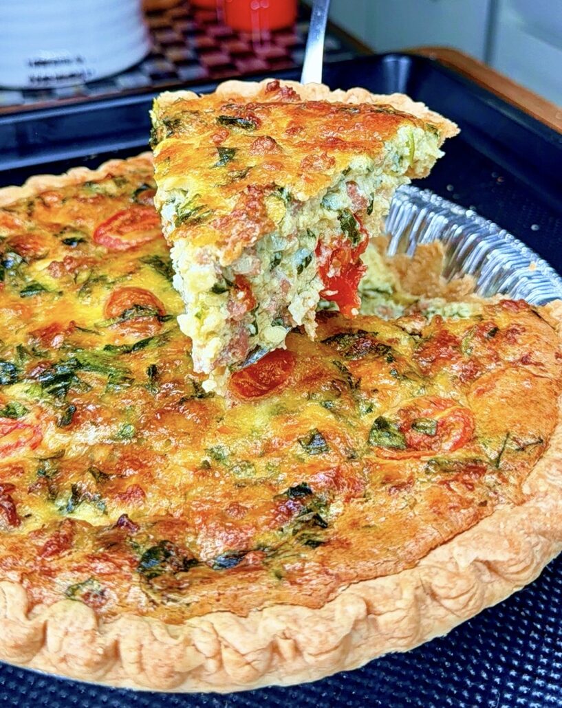 A close-up of a freshly baked quiche in a pie crust, filled with vegetables and herbs. One slice is being lifted out, showing a golden, cheesy, and colorful interior.