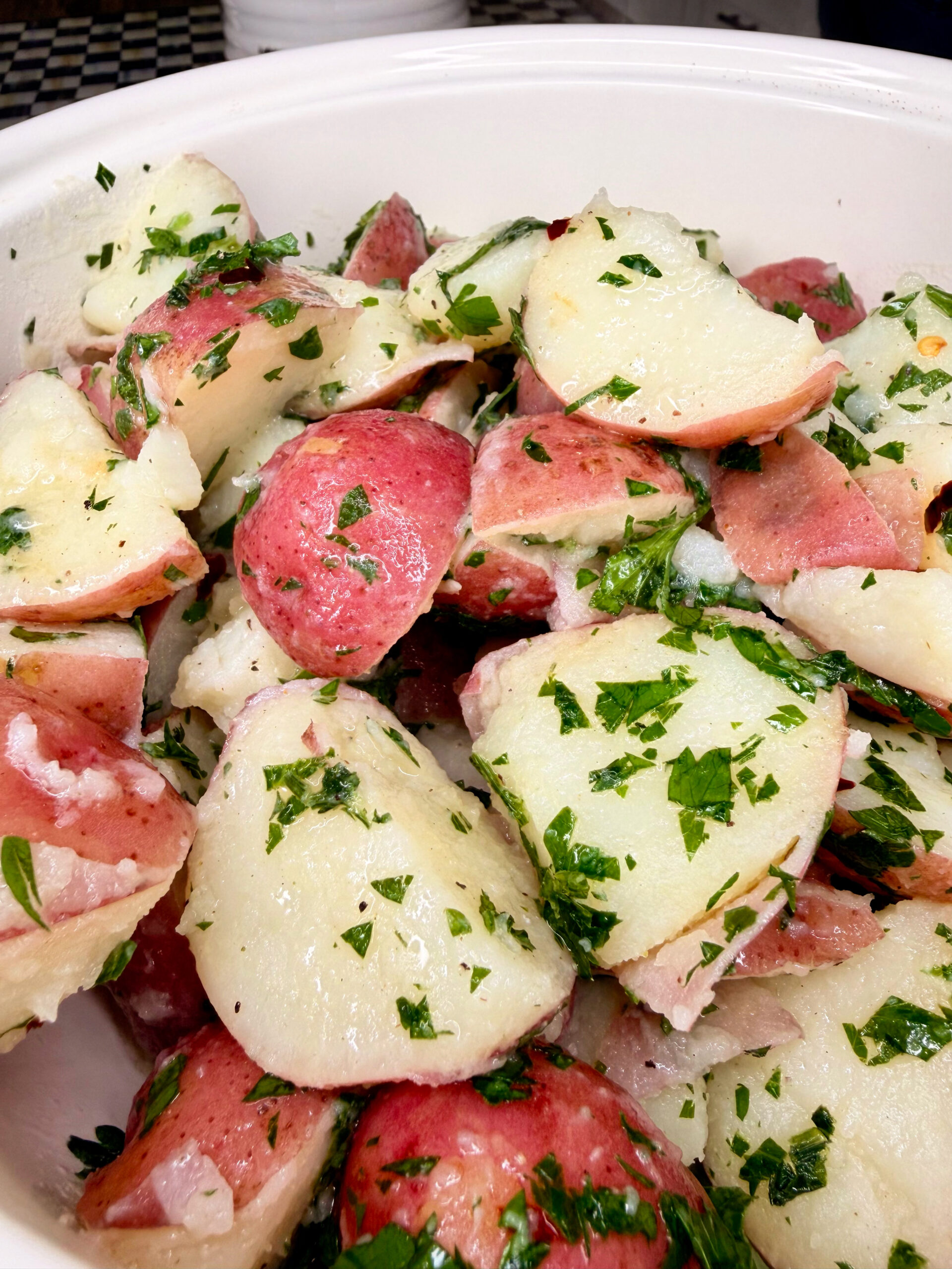 A close-up of a bowl filled with red potato salad, featuring sliced red potatoes mixed with fresh chopped parsley and a light dressing.