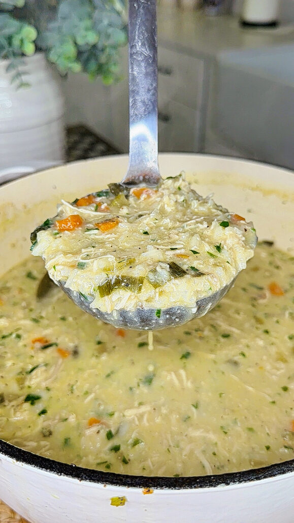 A close-up of a ladle holding a serving of creamy chicken and rice soup with vegetables, above a pot filled with the same soup in a kitchen setting.