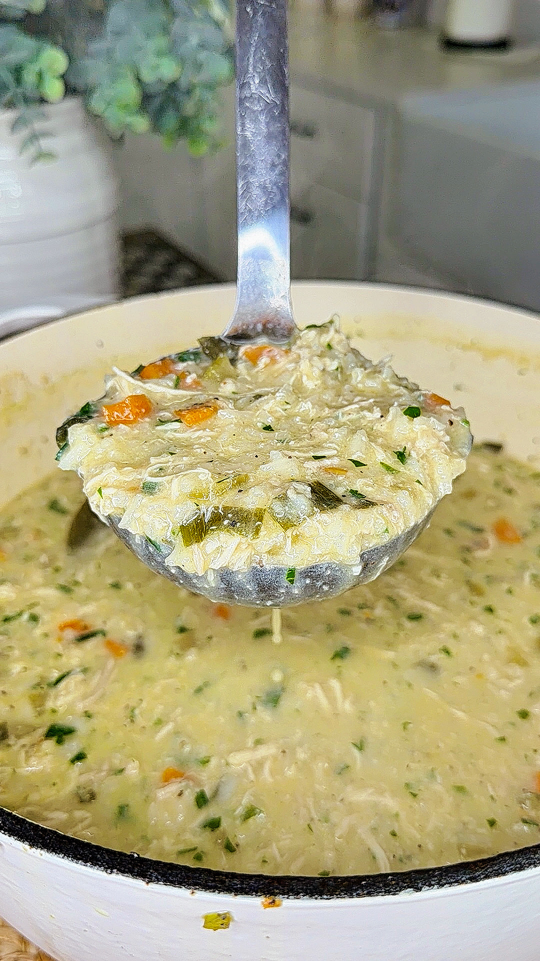 A close-up of a ladle holding a serving of creamy chicken and rice soup with vegetables, above a pot filled with the same soup in a kitchen setting.