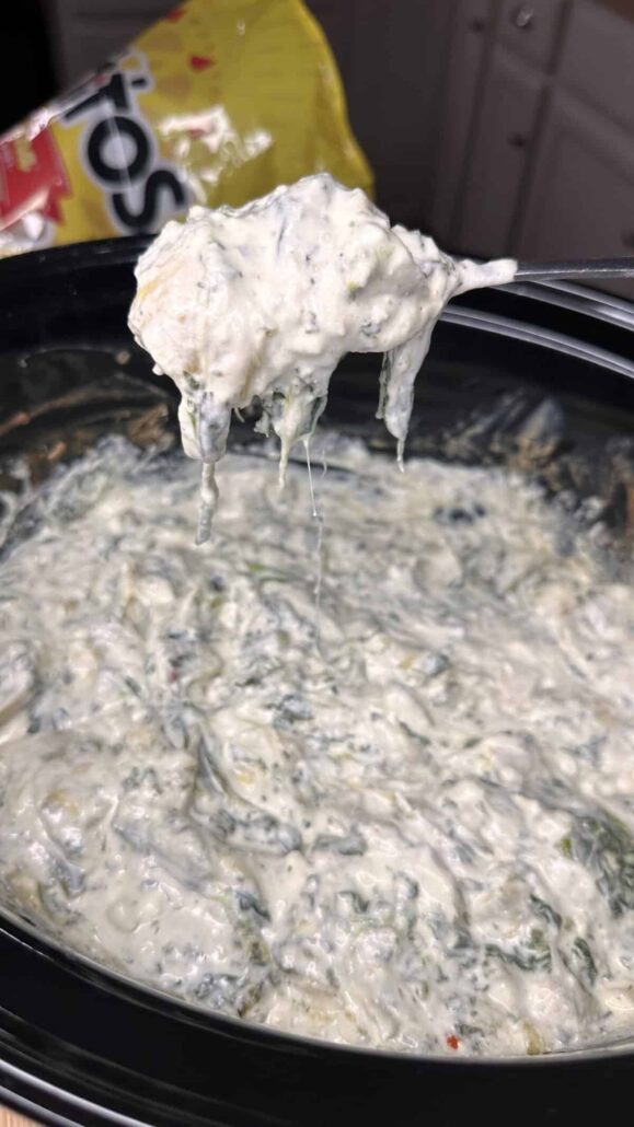 A spoon lifts a serving of creamy spinach and artichoke dip from a slow cooker, with a bag of tortilla chips in the background. The dip appears hot, cheesy, and gooey.