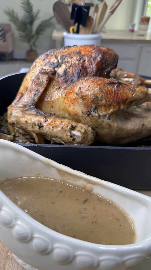 A roasted turkey with crispy, golden brown skin sits in a black roasting pan, with a white gravy boat filled with brown gravy in the foreground; kitchen utensils and plants are blurred in the background.