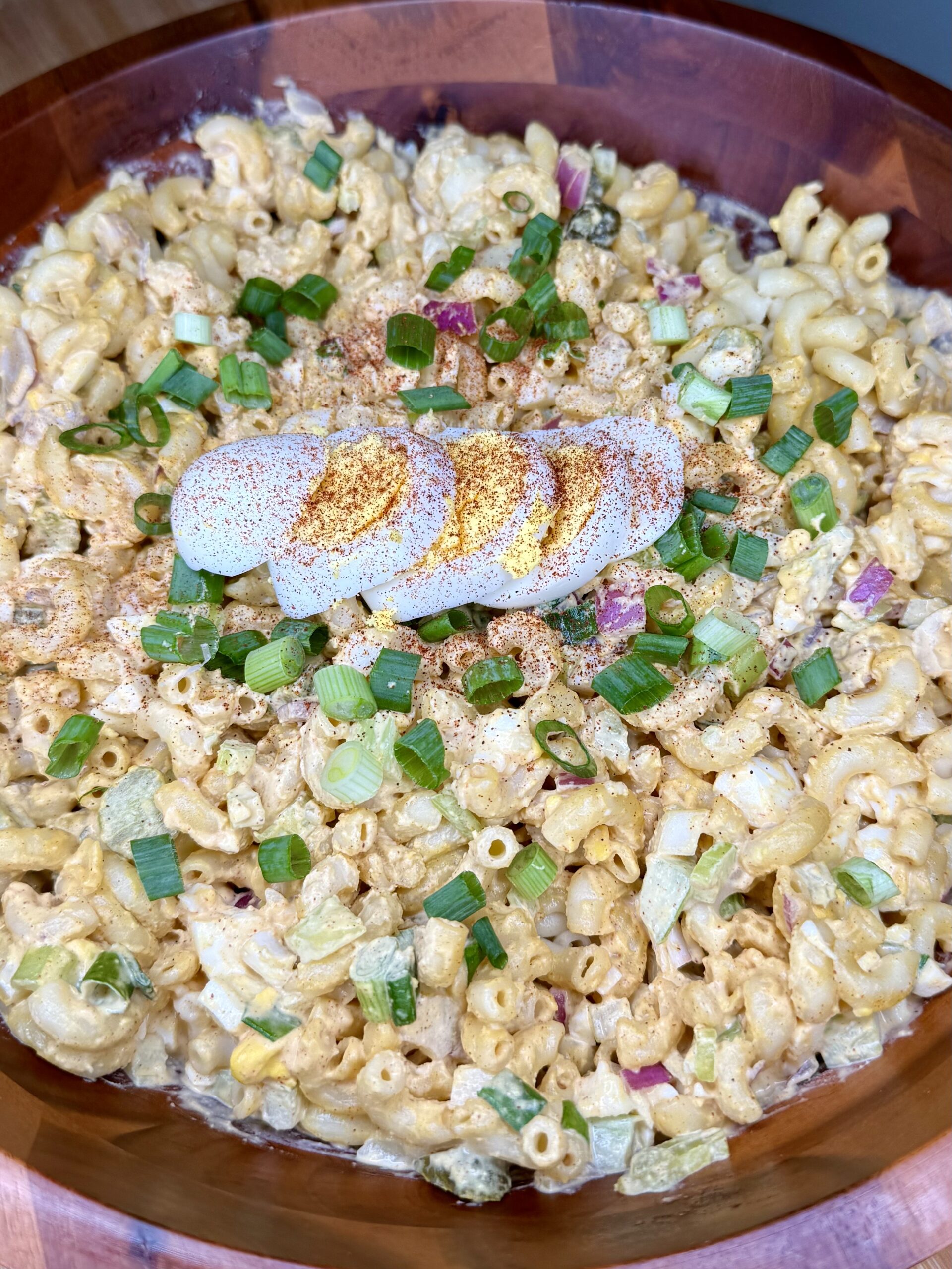 A wooden bowl filled with macaroni salad, topped with sliced hard-boiled eggs sprinkled with paprika, and garnished with chopped green onions.