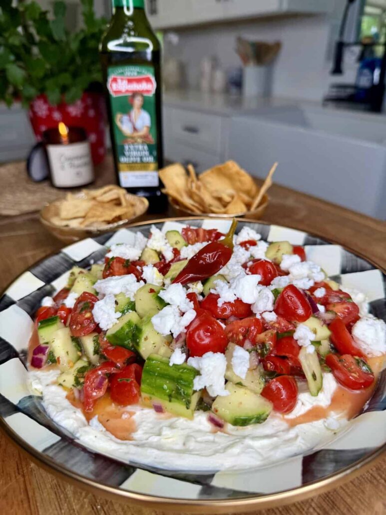 A plate of Mediterranean dip topped with chopped cucumbers, tomatoes, onions, and crumbled cheese sits on a table. A bowl of pita chips, a bottle of olive oil, and a lit candle are visible in the background.