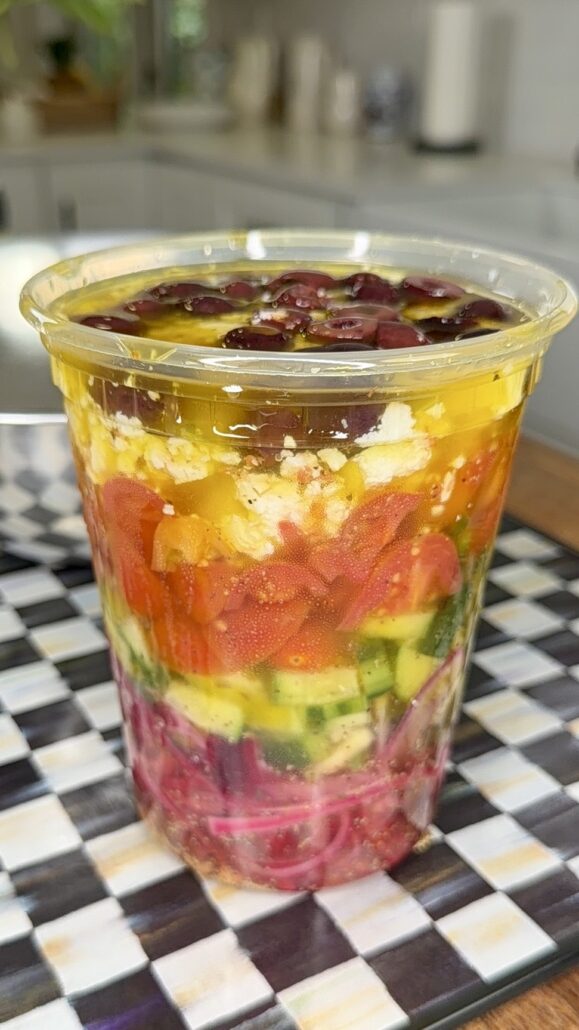 A clear plastic cup filled with colorful layers of chopped vegetables, cheese, and dressing, arranged in stripes, sitting on a checkered surface in a bright kitchen.