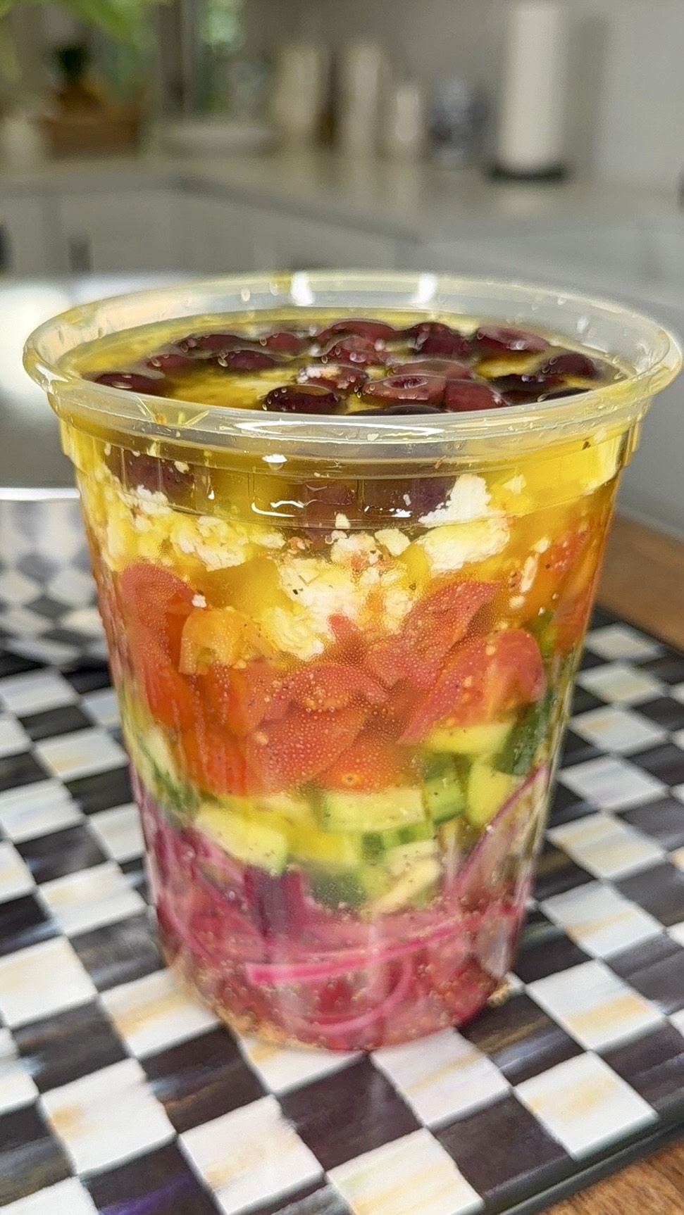 A clear plastic cup filled with colorful layers of chopped vegetables, cheese, and beans, sitting on a checkered surface in a bright kitchen.