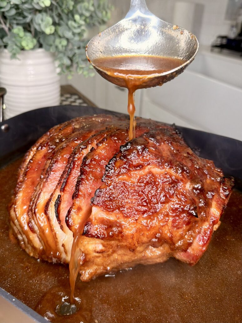 A close-up of a glazed, baked ham in a roasting pan as a hand pours brown sauce over it with a ladle. Steam rises from the ham, and a plant is visible in the blurred background.