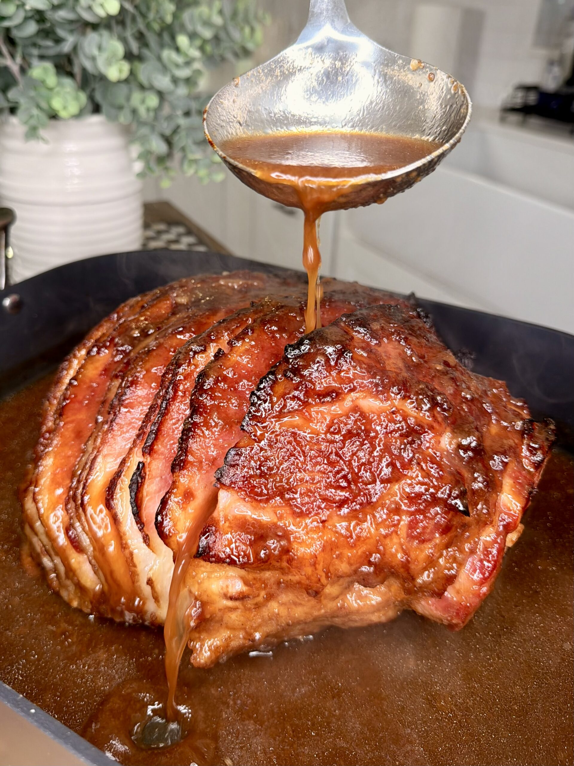 A close-up of a glazed, baked ham in a roasting pan as a hand pours brown sauce over it with a ladle. Steam rises from the ham, and a plant is visible in the blurred background.