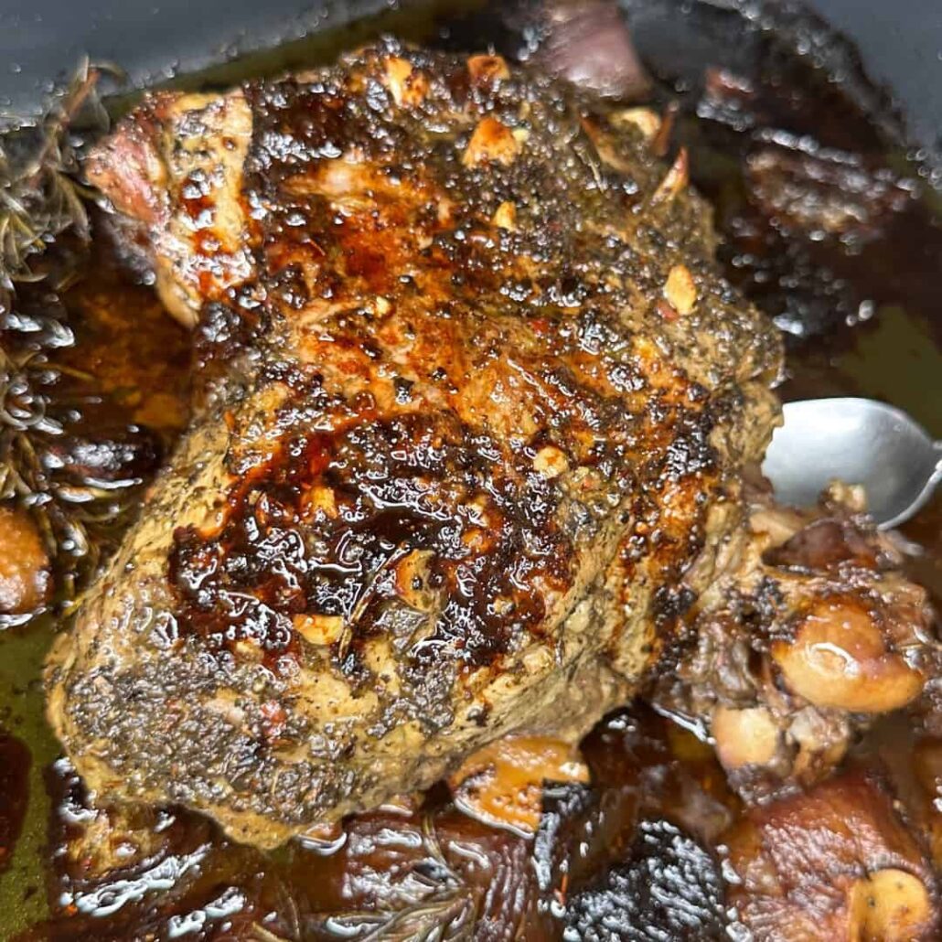 A close-up of a seasoned, roasted meat joint with a crispy, herb-crusted surface, surrounded by dark, caramelized juices and cooked vegetables, with a spoon partially visible on the right side.