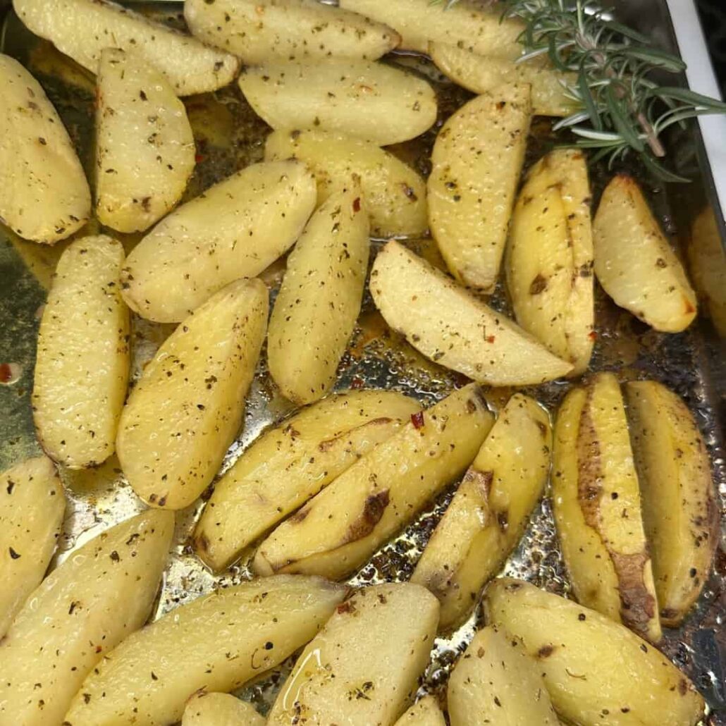 Golden roasted potato wedges sprinkled with herbs and spices on a baking tray, with a sprig of fresh rosemary in the corner.