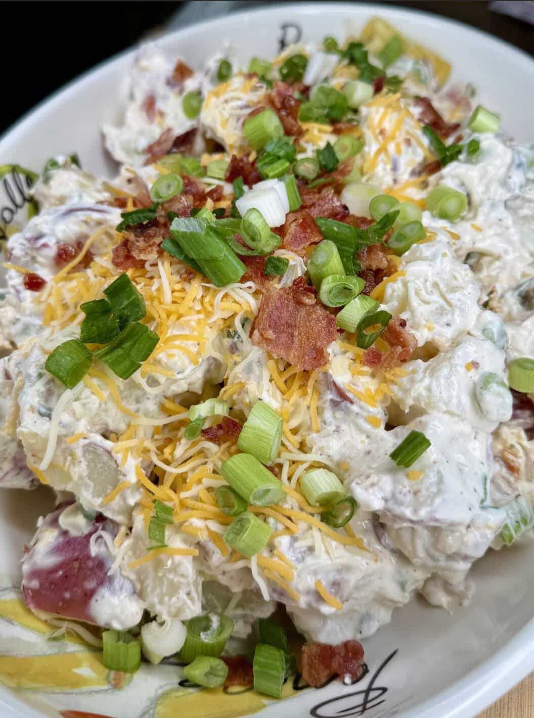 A close-up of a bowl filled with creamy potato salad, topped with shredded cheese, chopped green onions, and crispy bacon pieces.