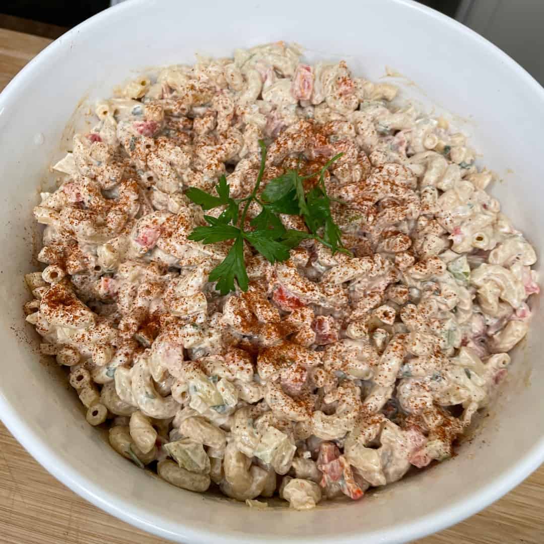 A bowl of macaroni salad with diced vegetables, coated in a creamy dressing, sprinkled with paprika, and garnished with fresh parsley on top.