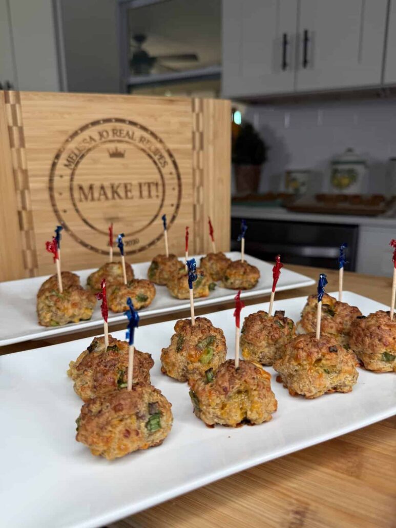 Plates of bite-sized meatball appetizers, each with a colored toothpick, are displayed on a wooden table in a kitchen. A wooden sign with engraved text stands in the background.