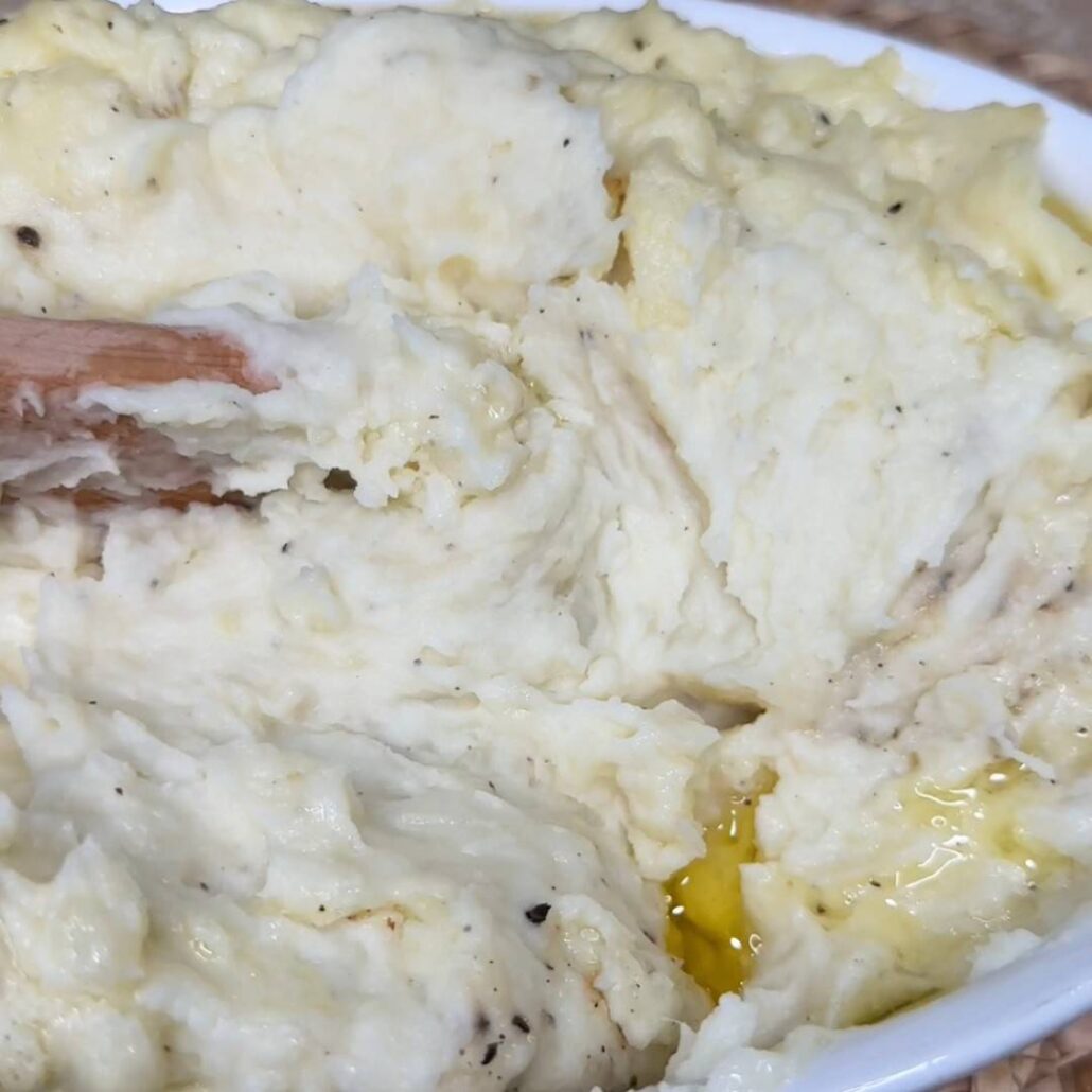 A close-up of creamy mashed potatoes with melted butter pooling on the surface and a wooden spoon partially buried in the mixture.