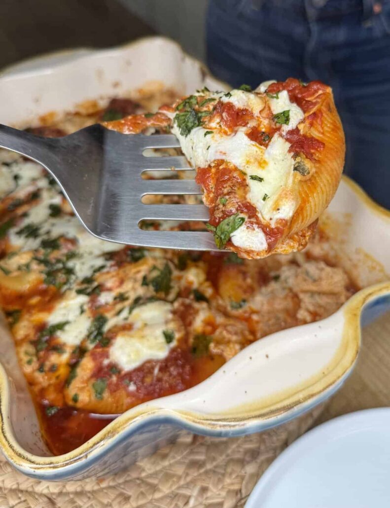 A metal spatula lifts a stuffed pasta shell topped with melted cheese, tomato sauce, and fresh herbs from a ceramic baking dish filled with more stuffed shells.