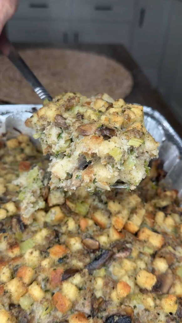 A close-up of a fork lifting a serving of homemade stuffing with pieces of bread, celery, and mushrooms from a foil baking tray. The background shows a kitchen countertop and cabinets.