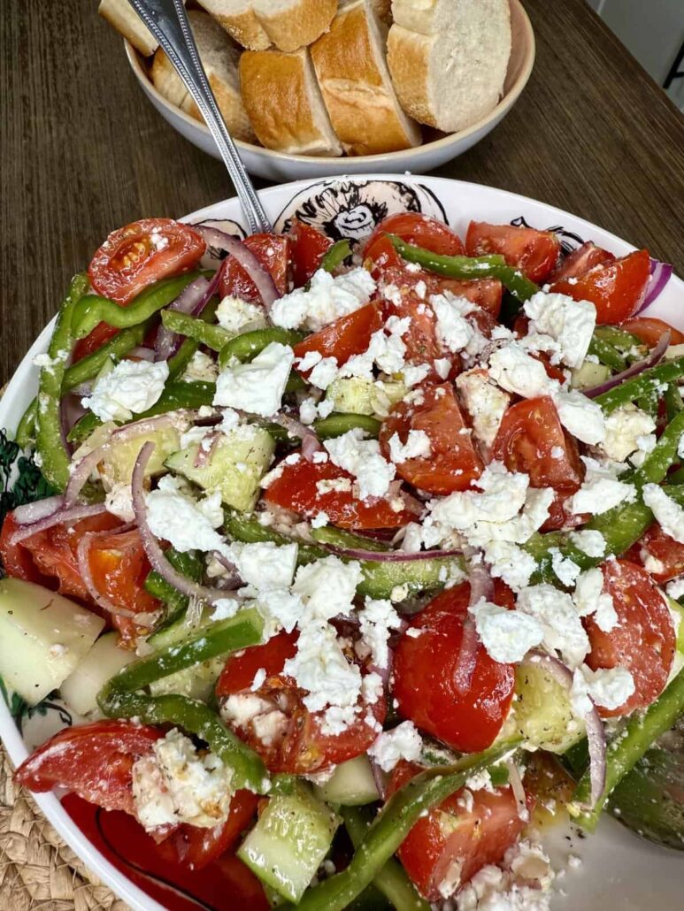 A bowl of Greek salad with tomatoes, cucumber, green peppers, red onions, and crumbled feta cheese, with a plate of sliced baguette bread in the background on a wooden table.