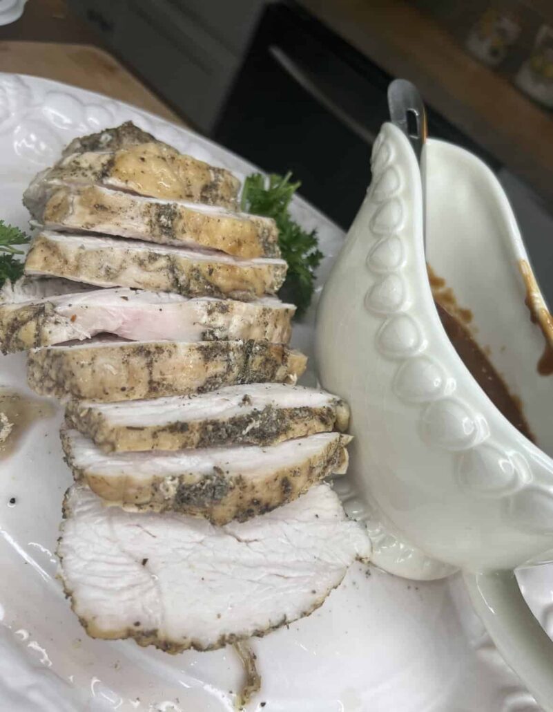 Sliced, herb-seasoned pork roast served on a white plate with a sprig of parsley, next to a white gravy boat filled with brown sauce.