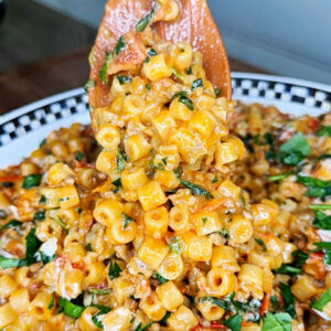 Close-up of a wooden spoon lifting a serving of ditalini pasta mixed with ground meat, tomato sauce, shredded carrots, and fresh spinach from a bowl, garnished with herbs and grated cheese.