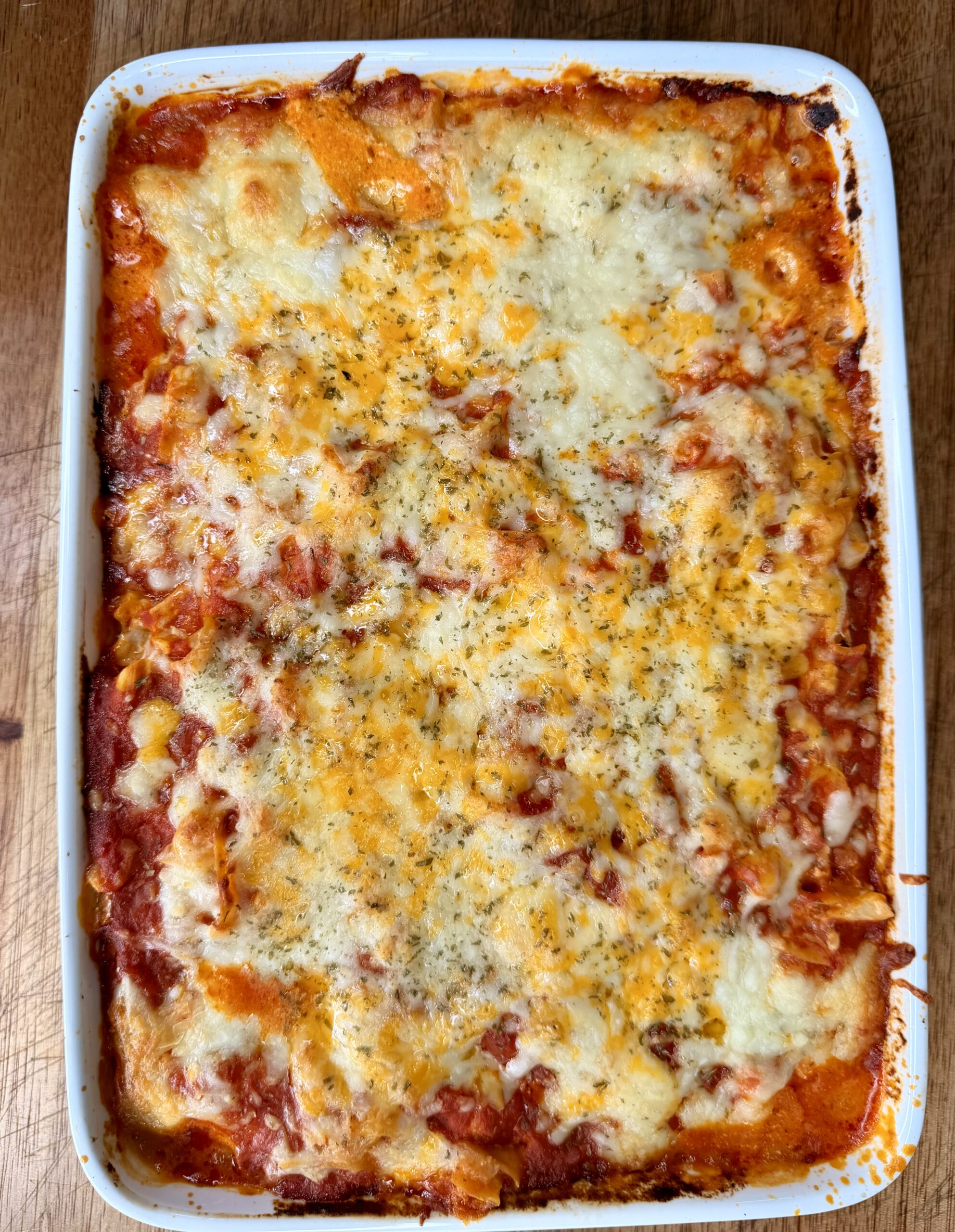 A baked casserole dish filled with golden, bubbly, and slightly browned cheesy lasagna, topped with melted cheese and herbs, sitting on a wooden surface.