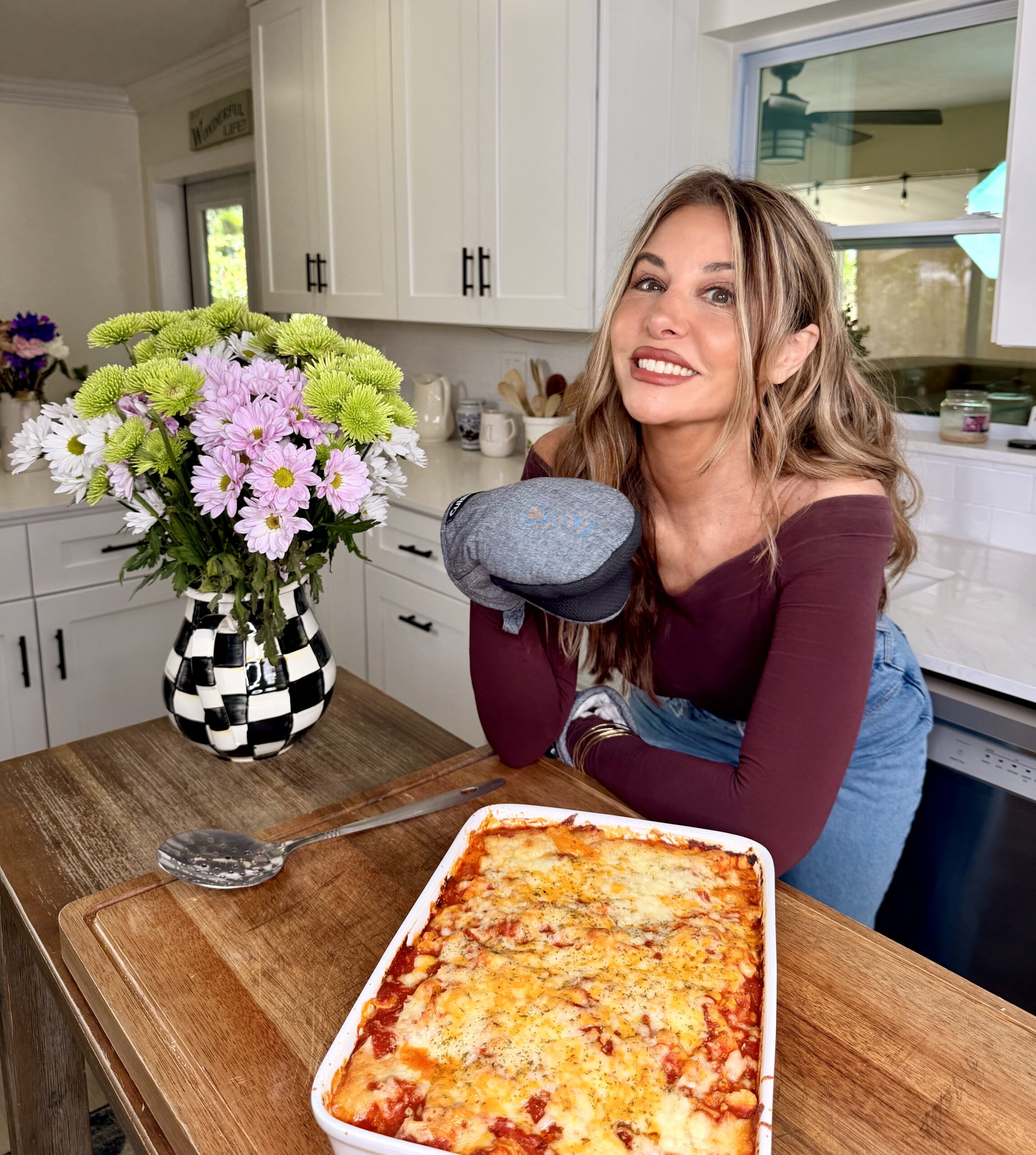 A smiling woman in a maroon top holds an oven mitt and stands in a kitchen next to a freshly baked lasagna on a wooden counter. A vase of purple and green flowers sits nearby.