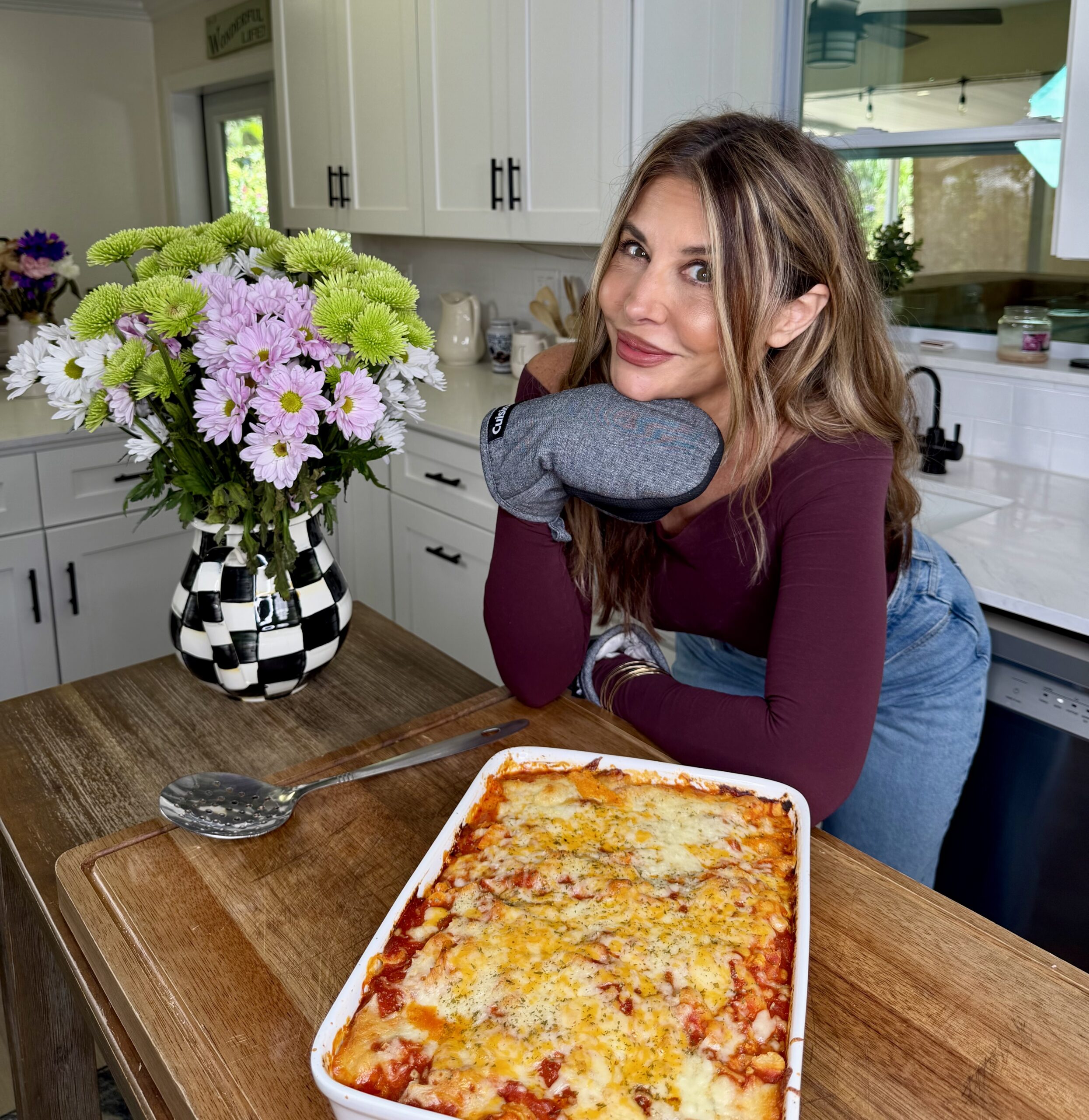 A woman in a kitchen, wearing an oven mitt and smiling, poses next to a freshly baked casserole on a wooden counter. A vase of purple and green flowers sits nearby.