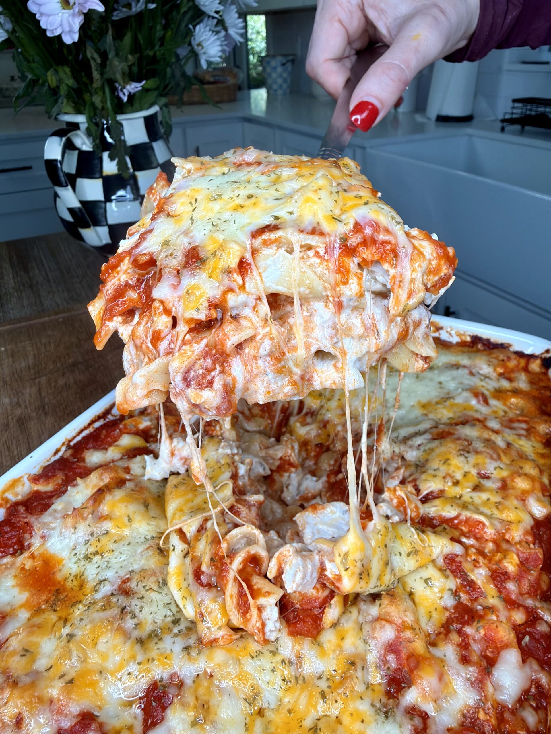 A hand lifts a gooey, cheesy slice of baked lasagna from a casserole dish, with melted cheese stretching out. A vase of flowers and a kitchen sink are visible in the background.