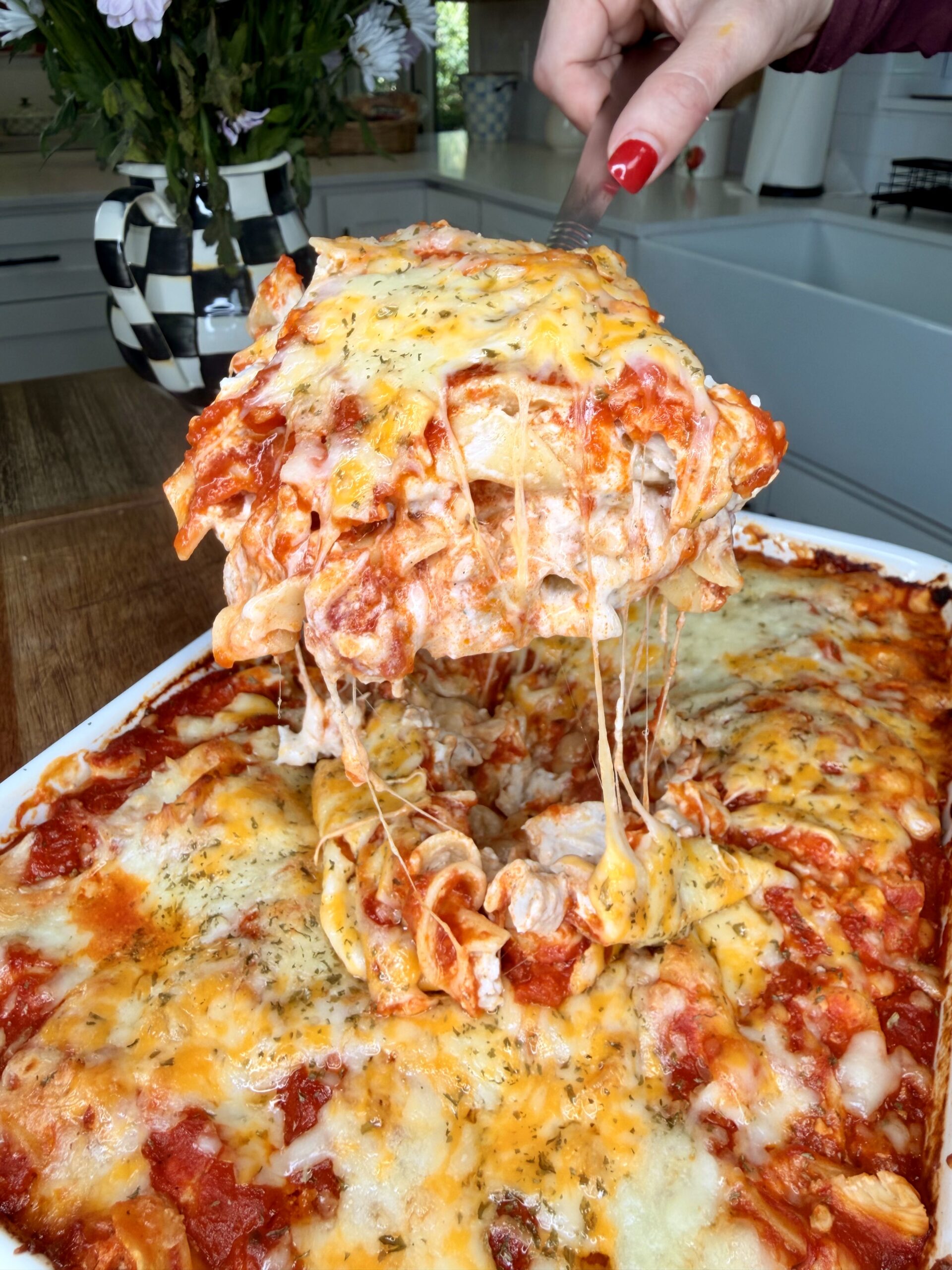 A hand lifts a cheesy, gooey slice of baked lasagna from a casserole dish, revealing layers of pasta, tomato sauce, and melted cheese in a bright kitchen setting.