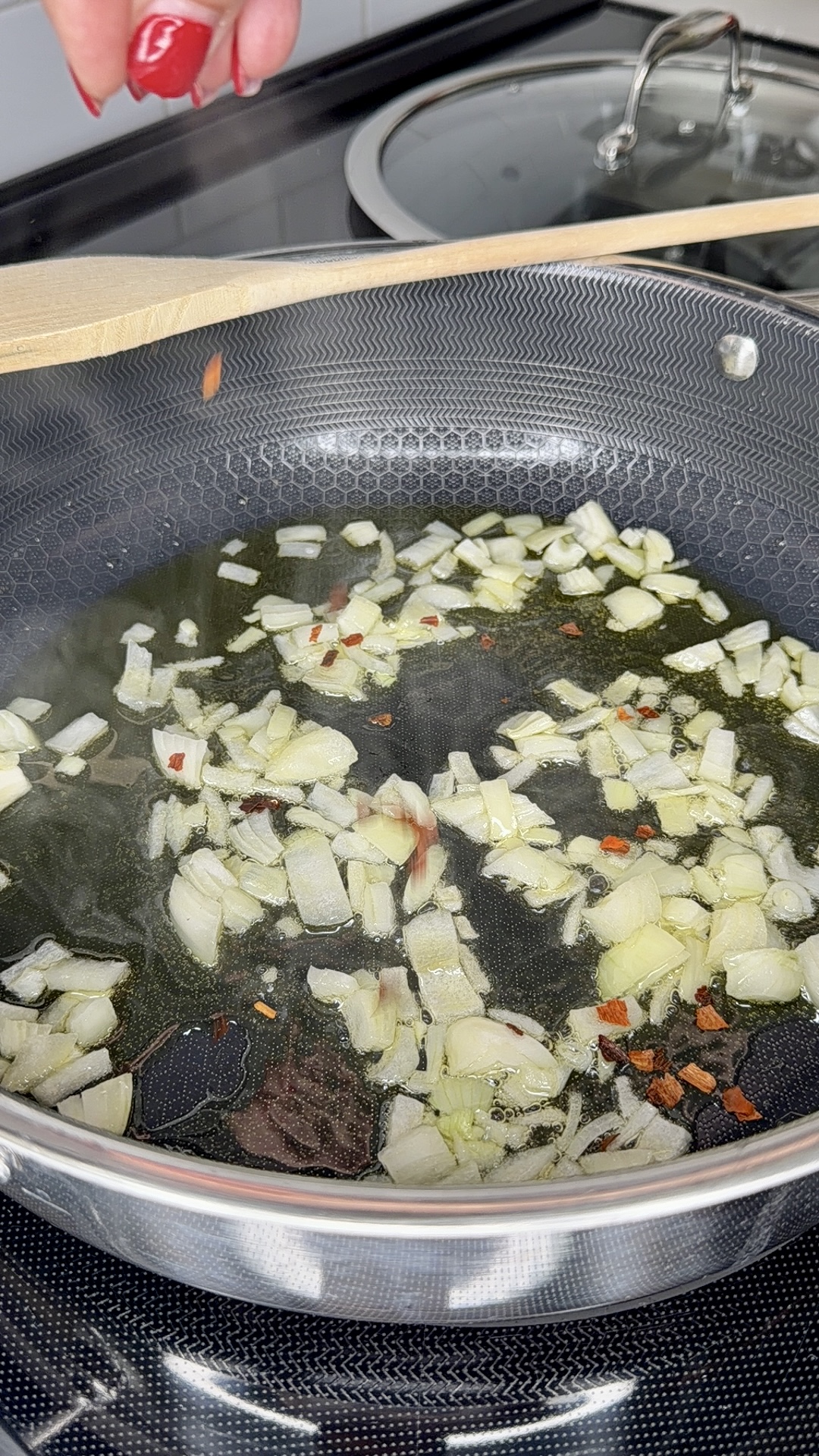 Chopped onions and red pepper flakes are sautéing in oil in a frying pan on a stove. A hand with red nails sprinkles more pepper flakes into the pan. A wooden spoon rests on the pans edge.