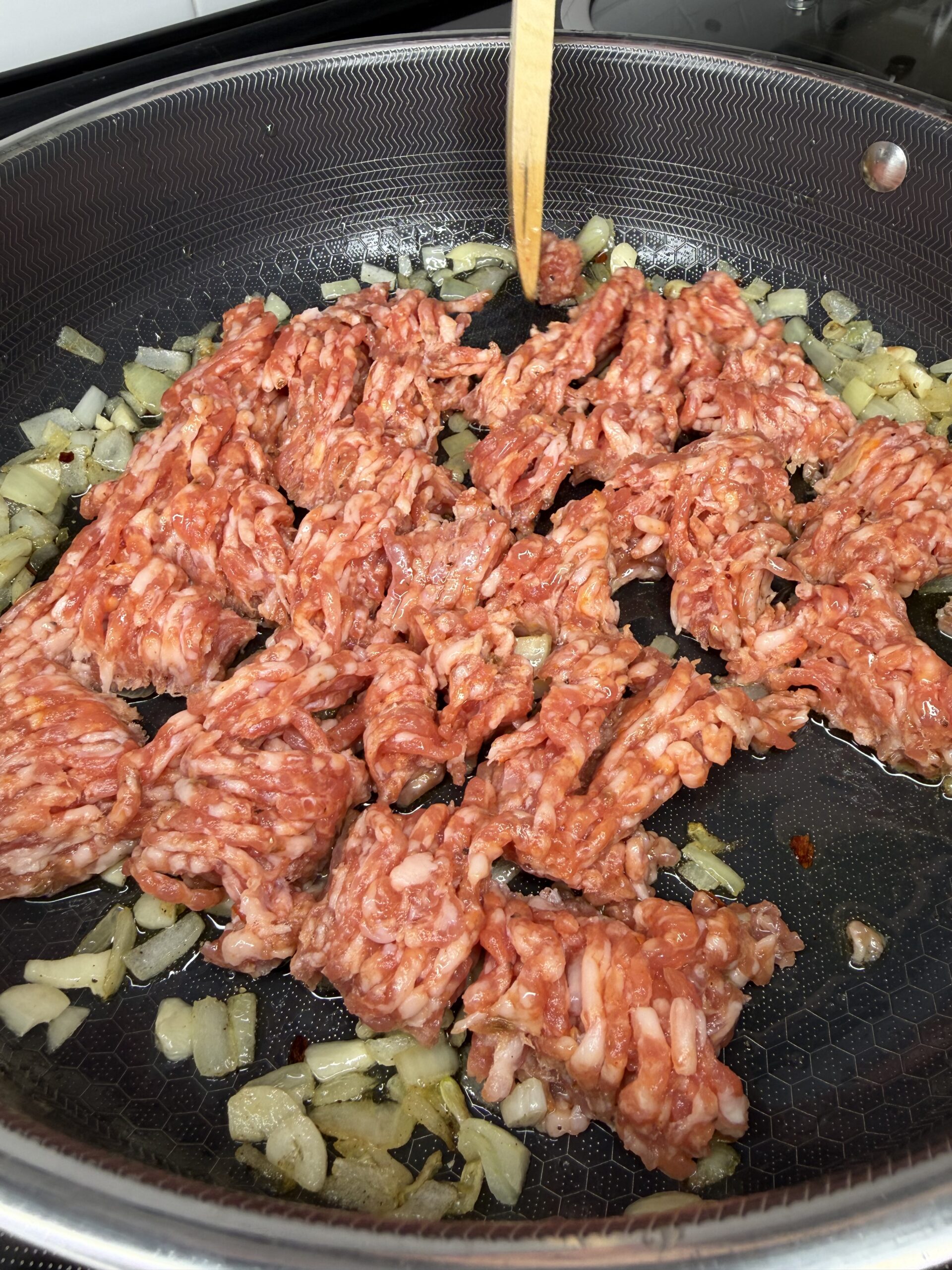 Raw ground meat and chopped onions are being cooked together in a frying pan, with a wooden utensil stirring the mixture.