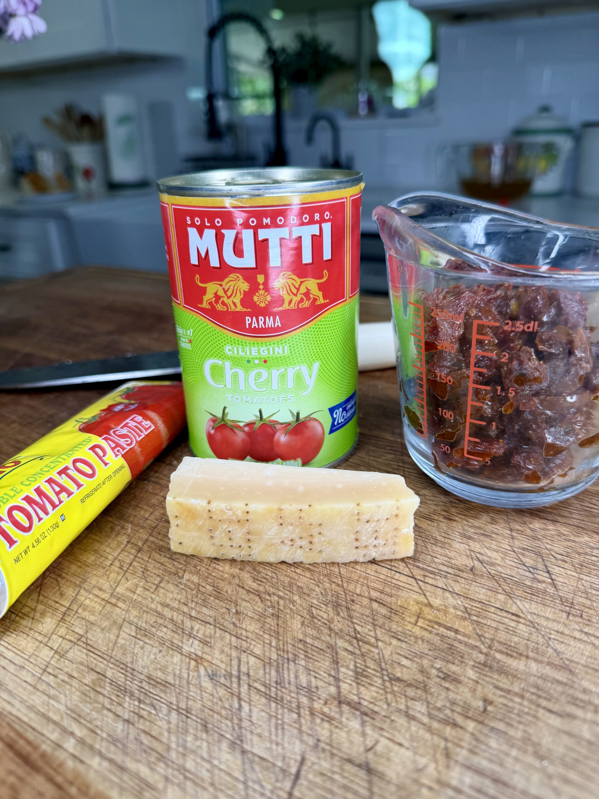 A can of Mutti cherry tomatoes, a glass measuring cup filled with sun-dried tomatoes, a tube of tomato paste, and a wedge of Parmesan cheese are arranged on a wooden cutting board in a kitchen.