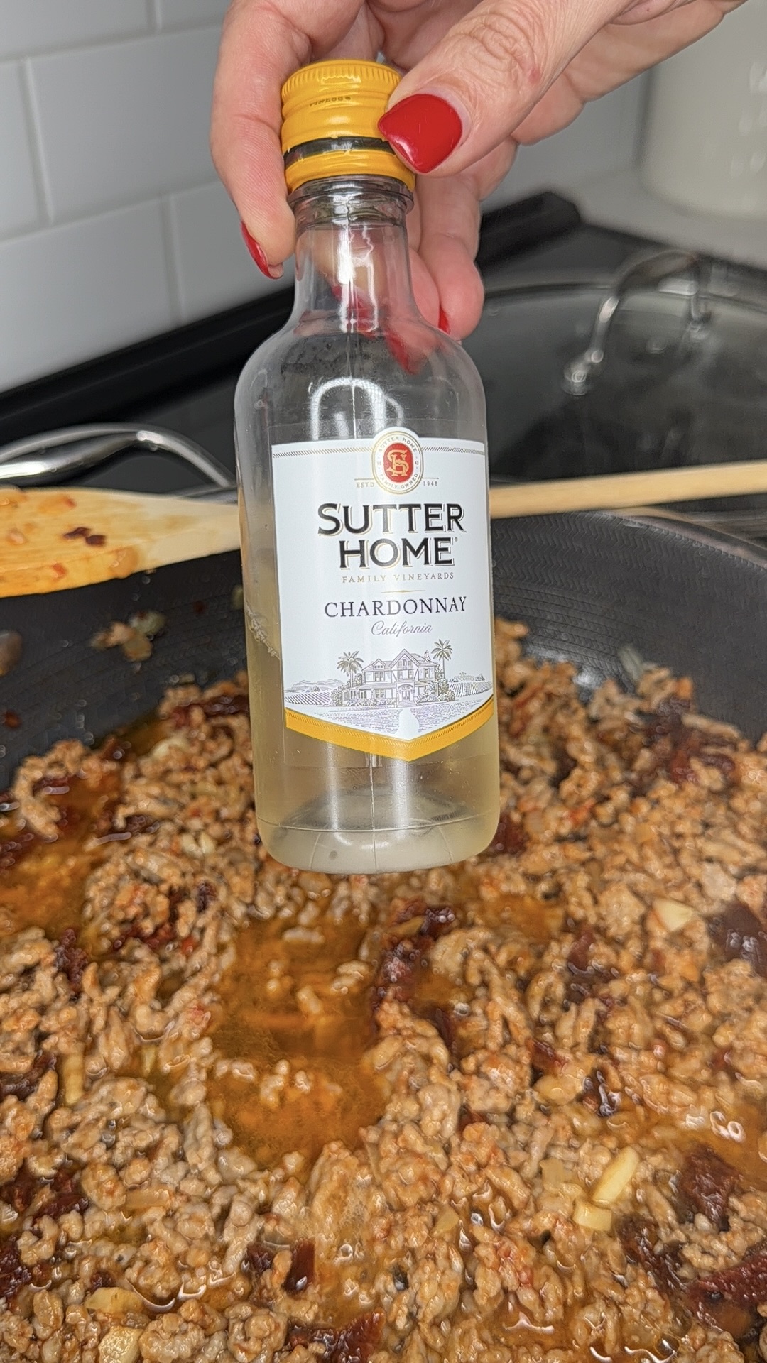 A hand with red nail polish holds a bottle of Sutter Home Chardonnay over a pan filled with browned ground meat cooking in sauce on a stovetop.