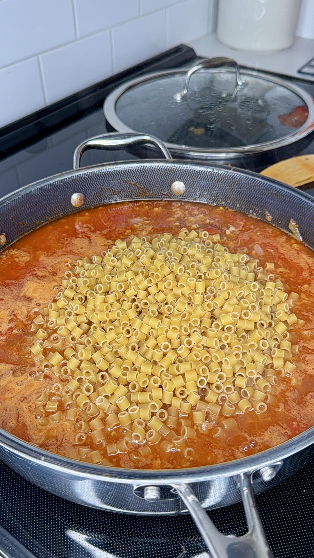 A large skillet filled with tomato sauce and uncooked ditalini pasta sits on a stove, with a glass lid and wooden spatula nearby.