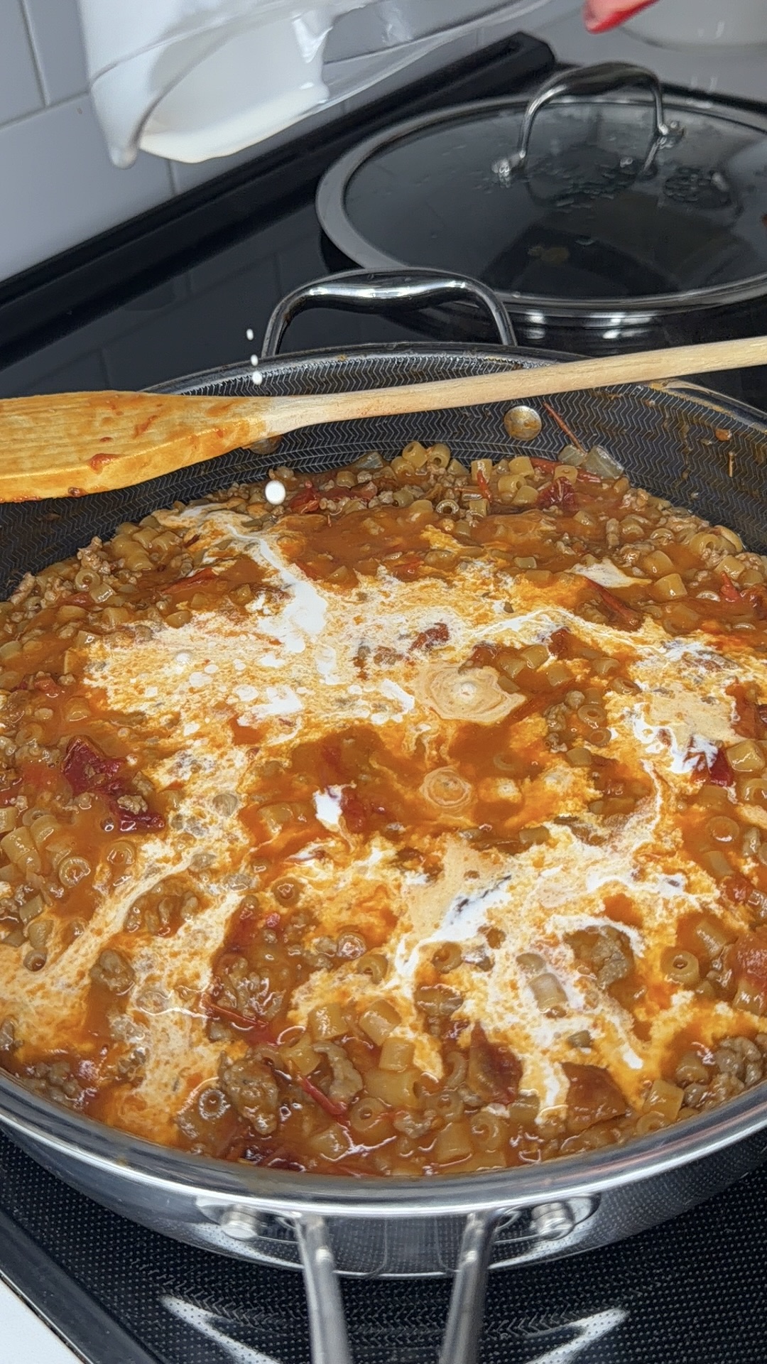 A pan on a stovetop contains a tomato-based lentil stew with a swirl of cream on top. A wooden spoon rests on the edge of the pan, and a pot with a glass lid is visible in the background.