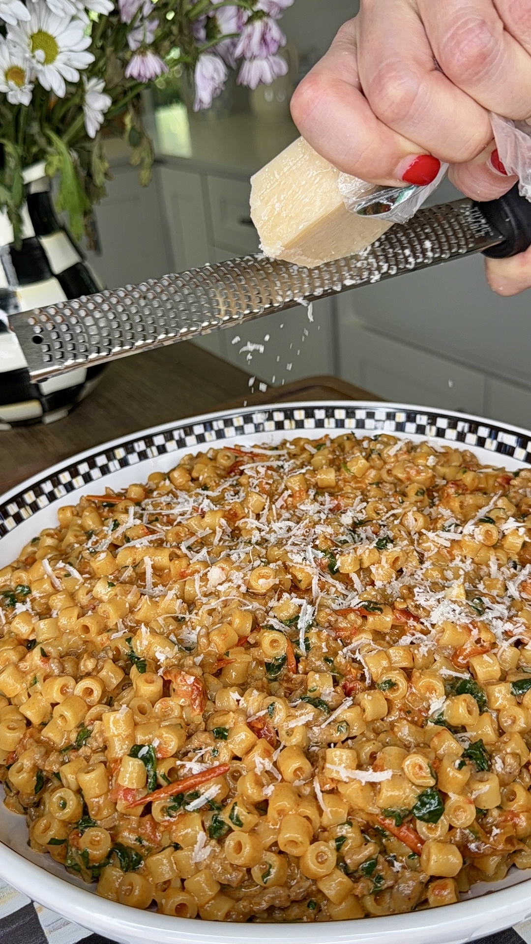 A hand grates a block of cheese over a bowl of cooked pasta with herbs and tomatoes. The pasta sits in a decorative black-and-white dish. A bouquet of flowers is visible in the blurred background.