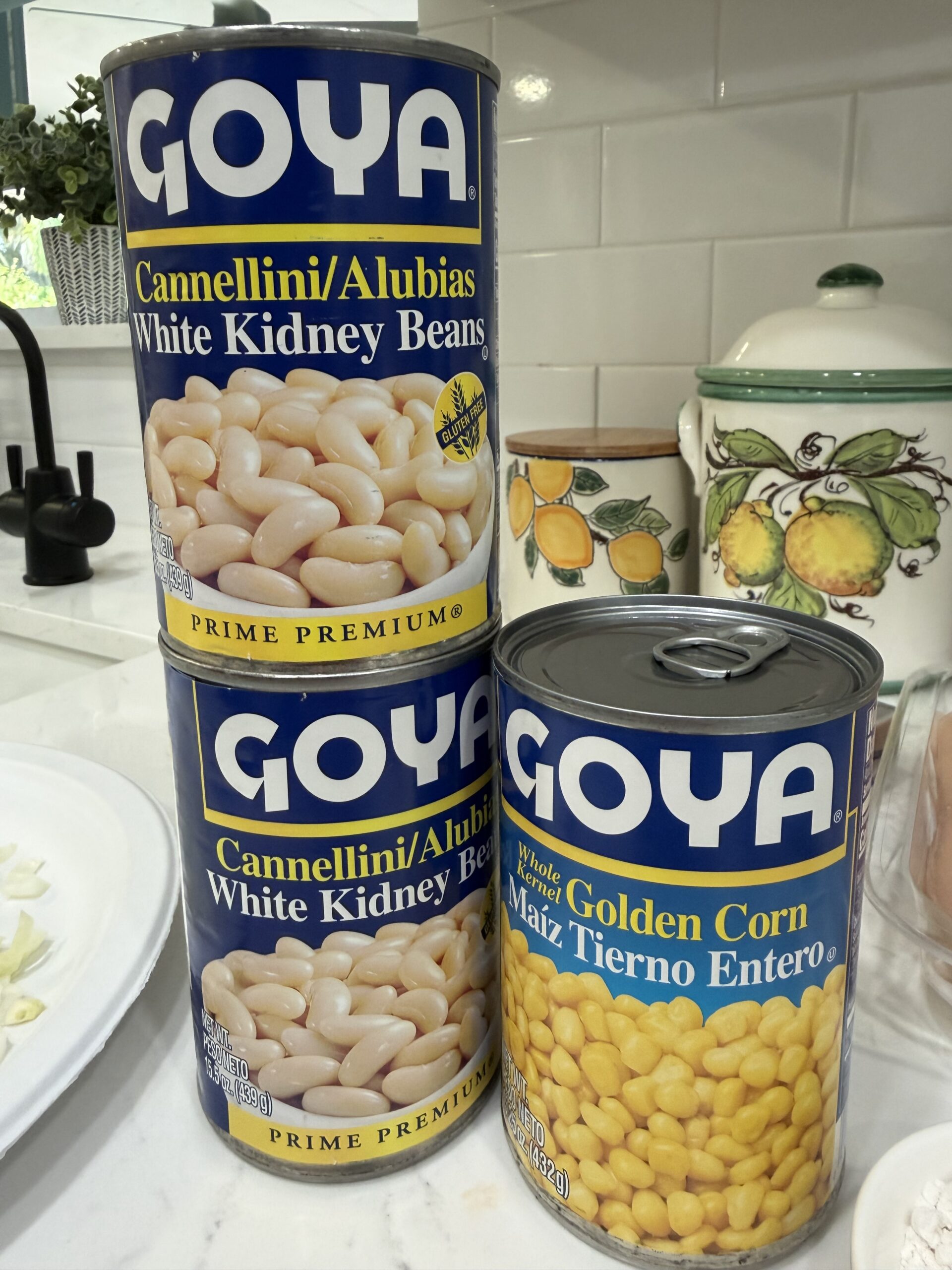 Three cans of Goya brand food on a kitchen counter: two cans of white kidney beans stacked on top of each other, and one can of whole golden corn beside them. A decorative lemon container is in the background.