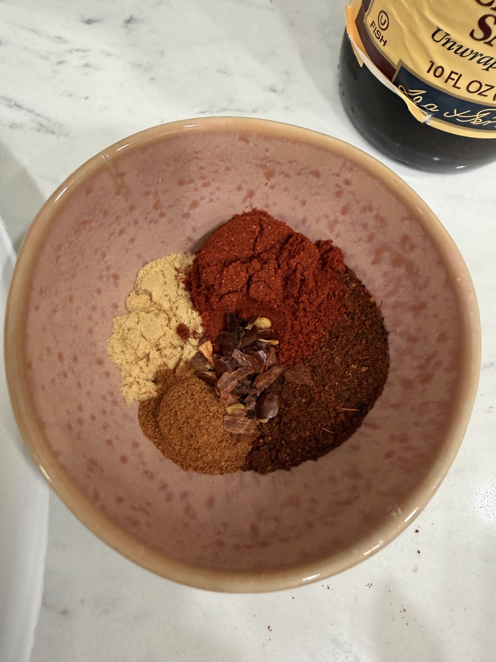 A pink bowl on a marble countertop contains small piles of ground ginger, paprika, chili powder, cinnamon, and red pepper flakes. A bottle of syrup is partially visible in the corner.
