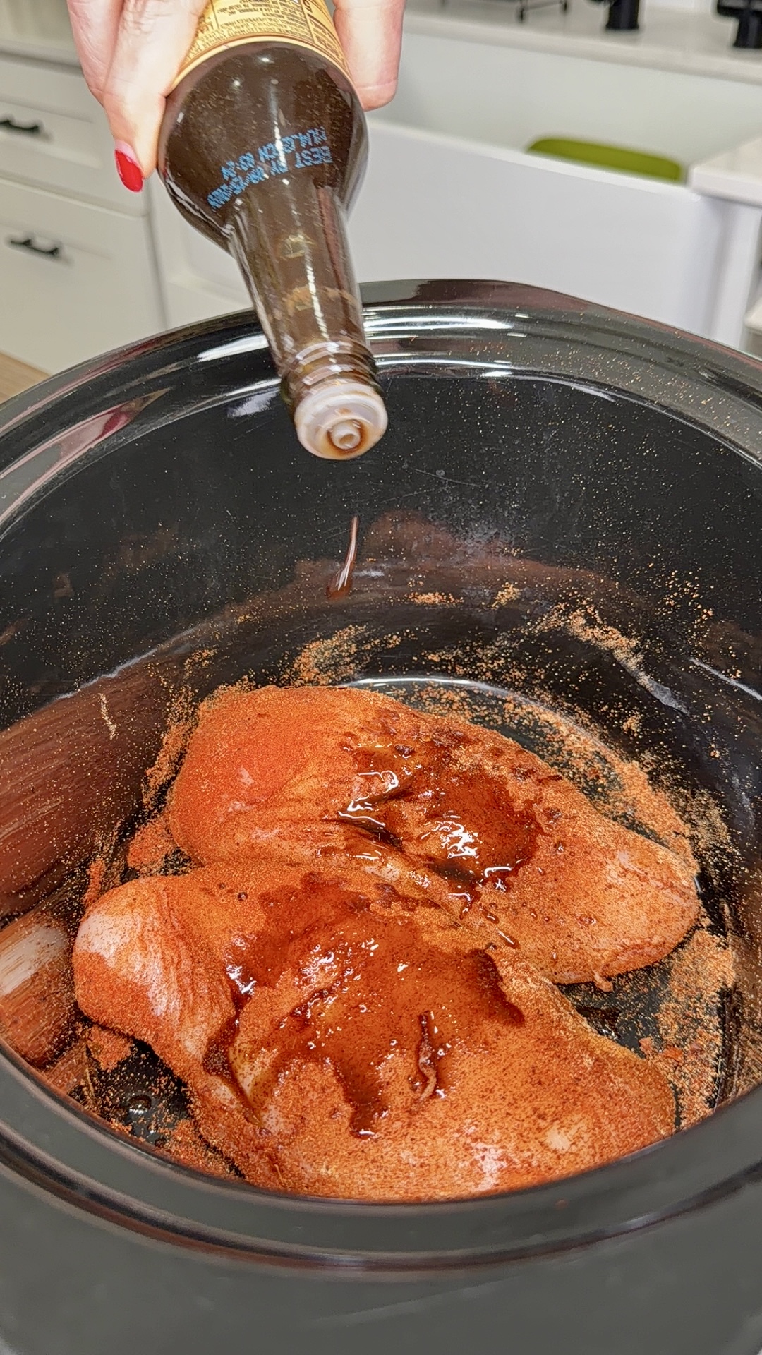 A hand pours a brown liquid from a bottle onto seasoned raw chicken breasts in a black slow cooker.