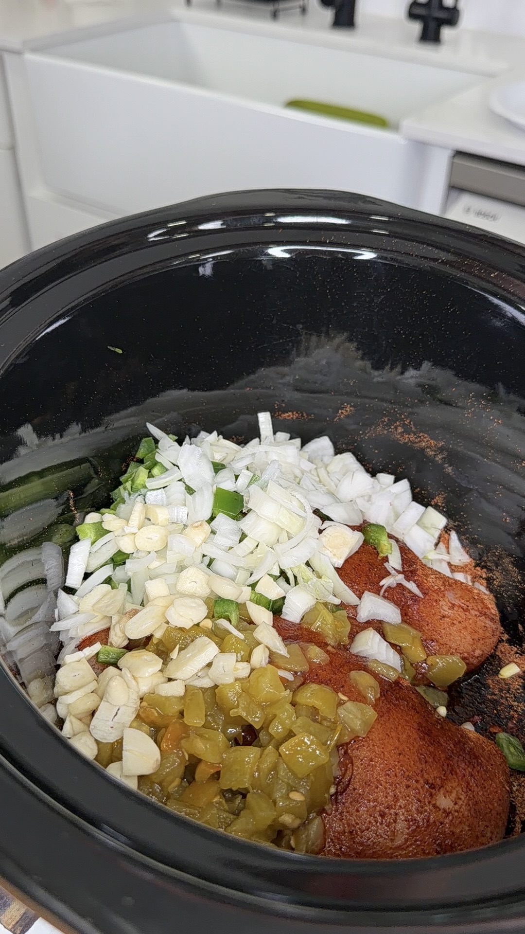 A slow cooker filled with chopped onions, sliced garlic, diced green chilies, green herbs, and red spices, ready to be mixed and cooked. A white kitchen sink is visible in the background.