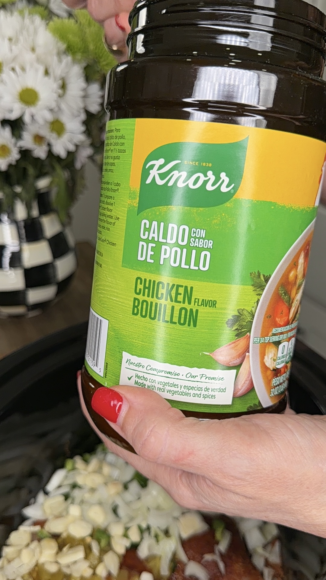 A hand with red nail polish holds a jar of Knorr Chicken Bouillon powder above a slow cooker filled with sliced garlic and onions. A bouquet of flowers is visible in the background.