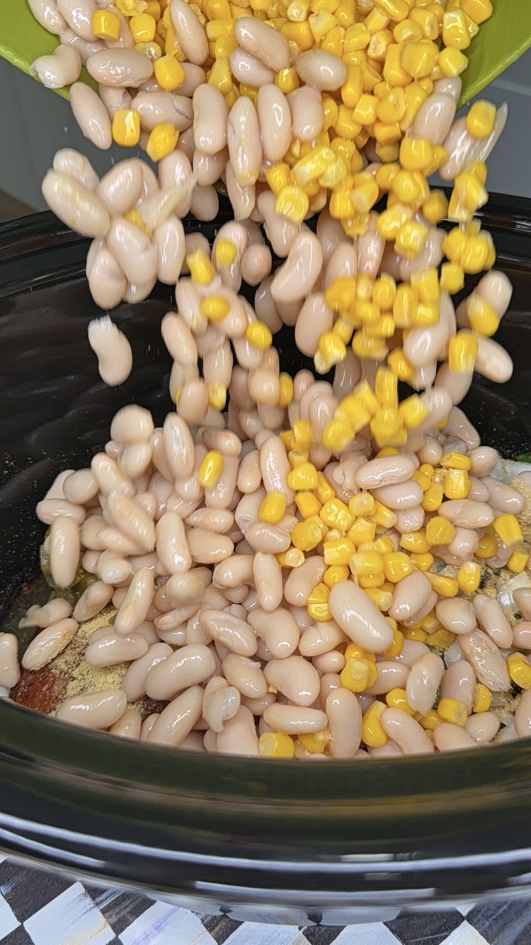 Canned white beans and yellow corn are being poured from a bowl into a black slow cooker with other visible seasonings at the bottom.