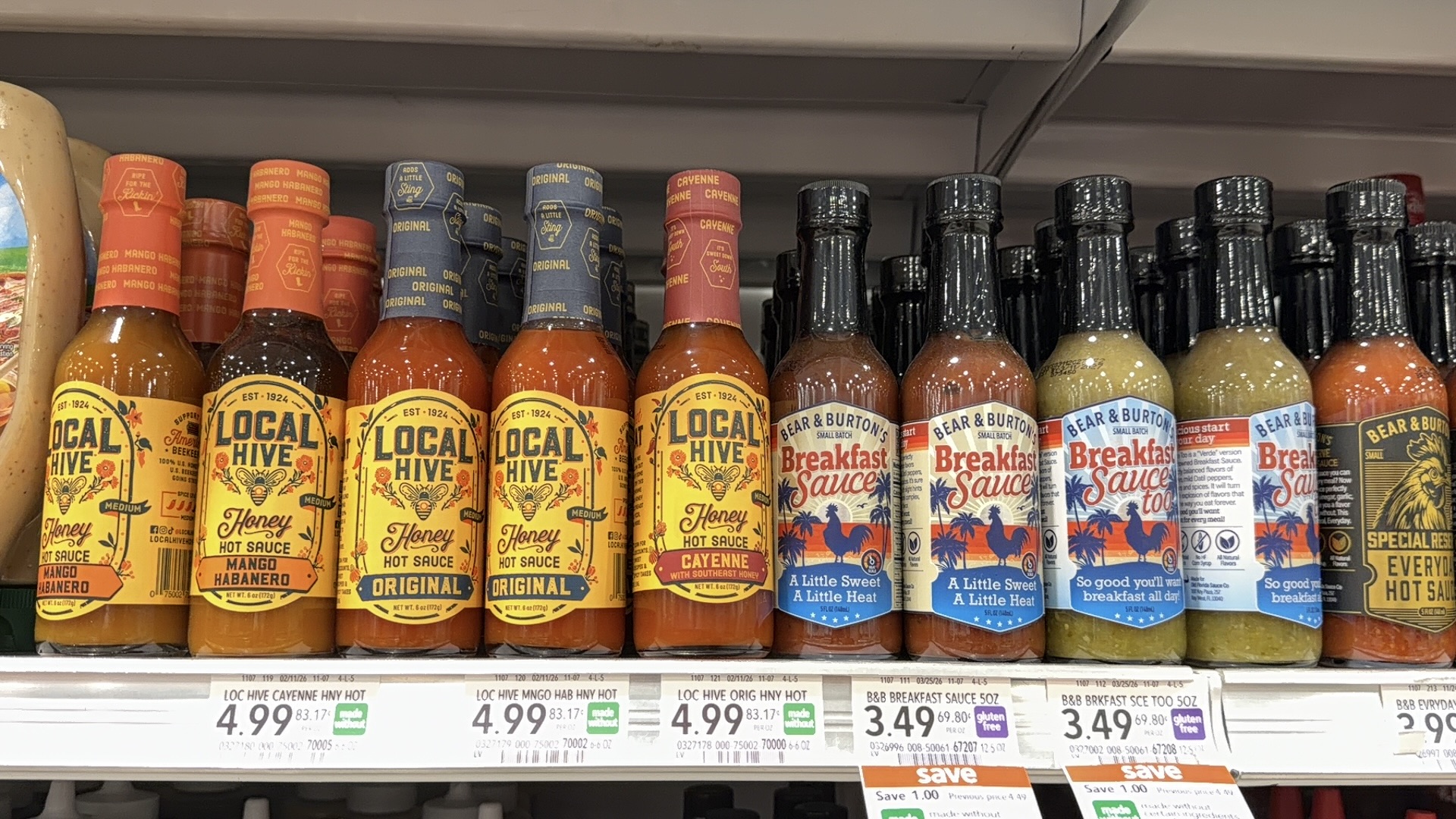 Bottles of Local Hive honey hot sauce and Bear & Burtons Breakfast Sauce varieties are displayed on a grocery store shelf, each with visible colorful labels and price tags below.