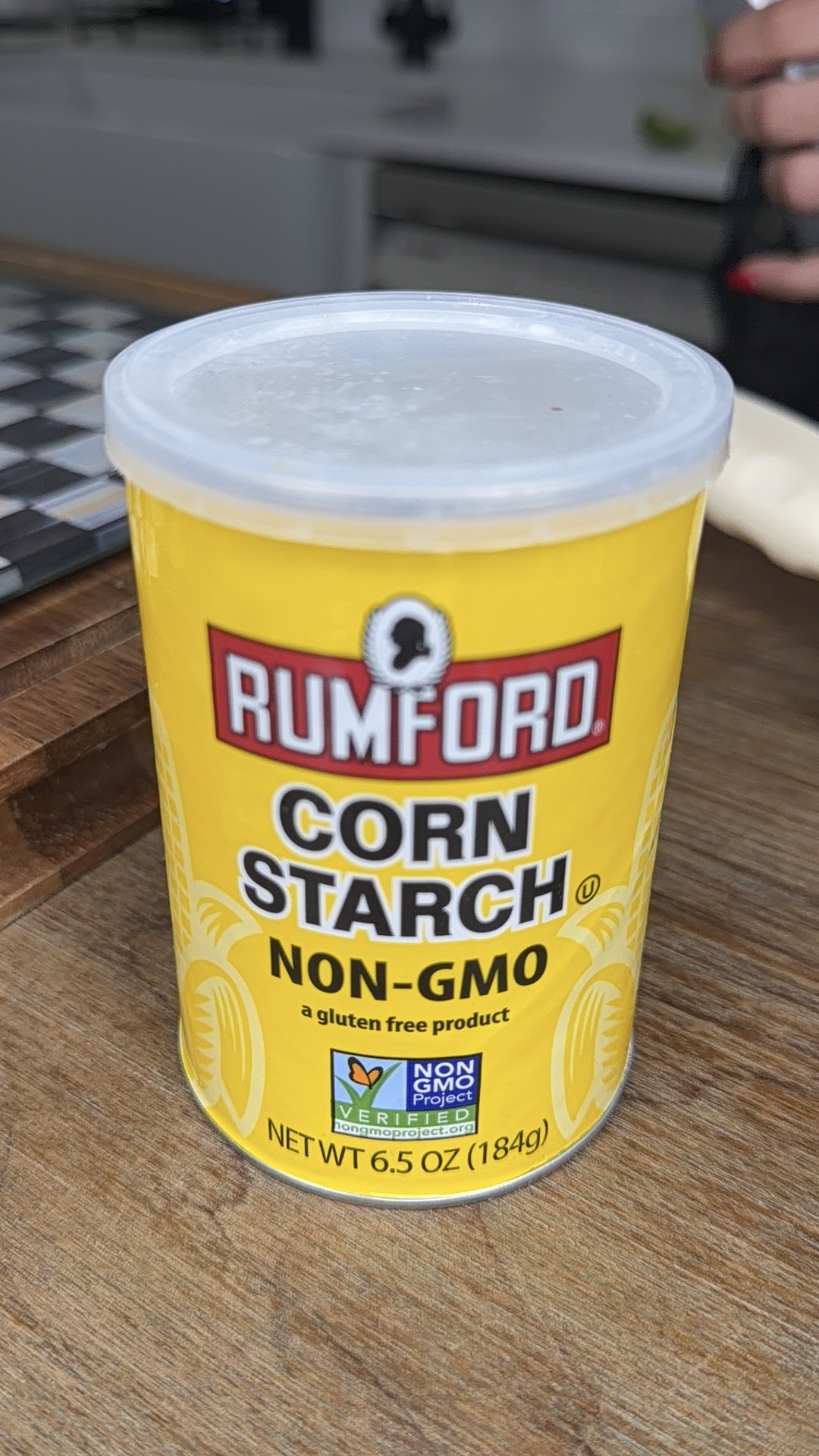 A yellow container of Rumford Corn Starch labeled NON-GMO, a gluten free product, and NET WT 6.5 OZ (184g), sits on a wooden surface with part of a kitchen visible in the background.