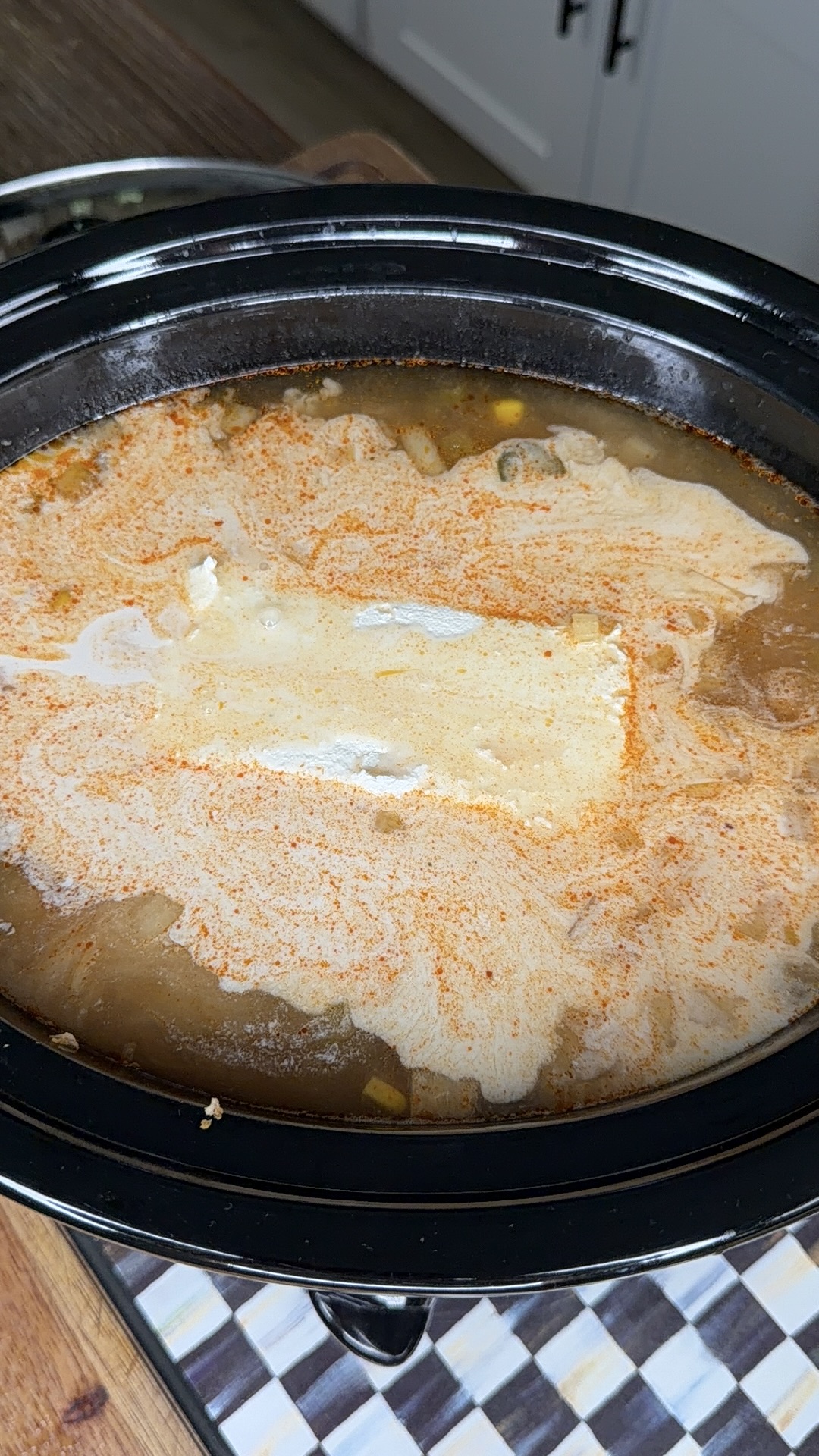 A slow cooker filled with soup, topped with a swirl of cream and sprinkled with reddish seasoning, sits on a wooden counter next to a checkered cloth.