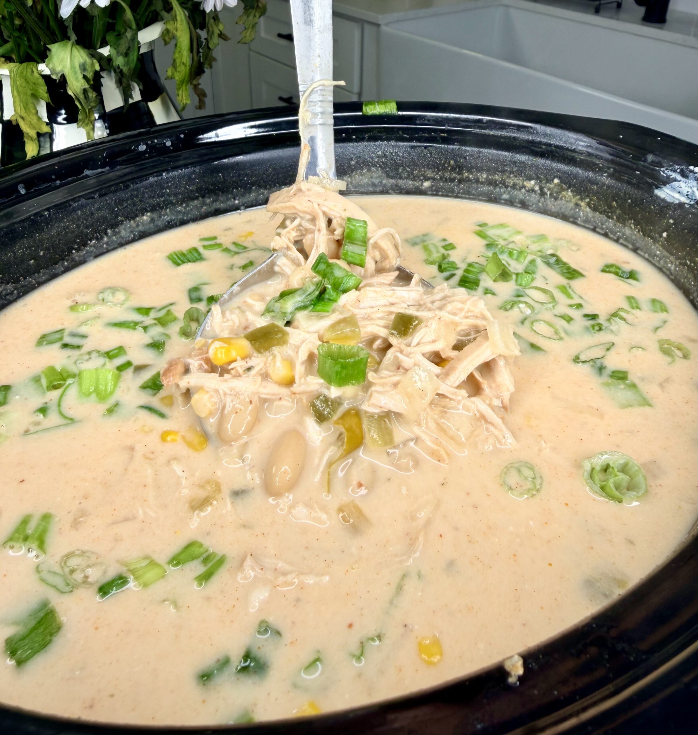 A close-up of creamy soup in a slow cooker, filled with shredded chicken, corn, white beans, green chilies, and topped with chopped green onions. A ladle is submerged in the soup, ready to serve.