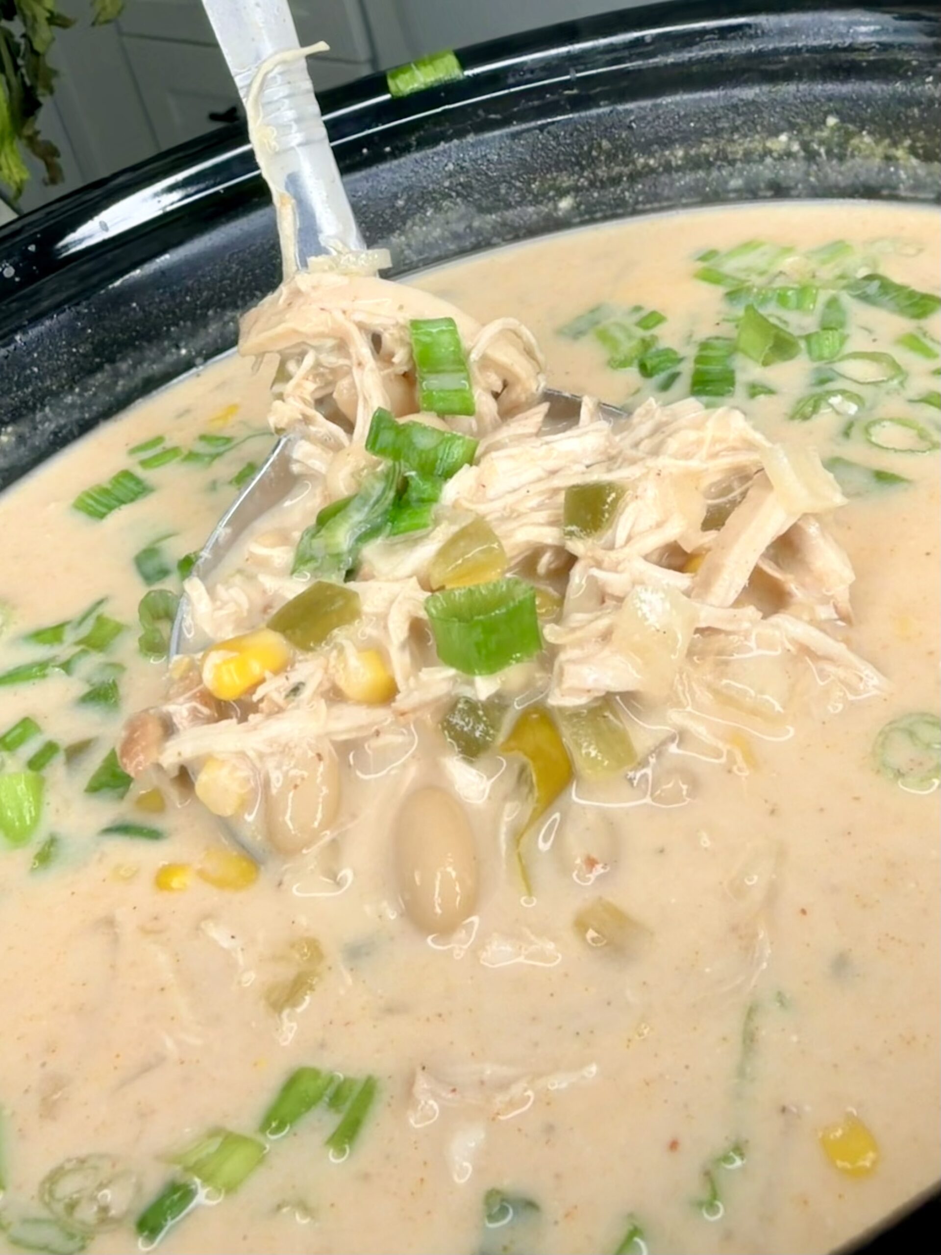 A creamy white chicken chili in a slow cooker, filled with shredded chicken, white beans, corn, and chopped green onions, with a spoon lifting a portion from the soup.