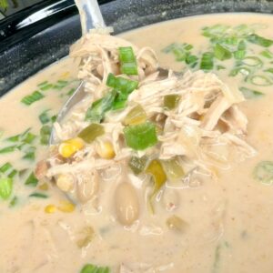 A creamy white chicken chili in a slow cooker, featuring shredded chicken, white beans, corn, diced green chilies, and chopped green onions, with a ladle scooping up a hearty serving.