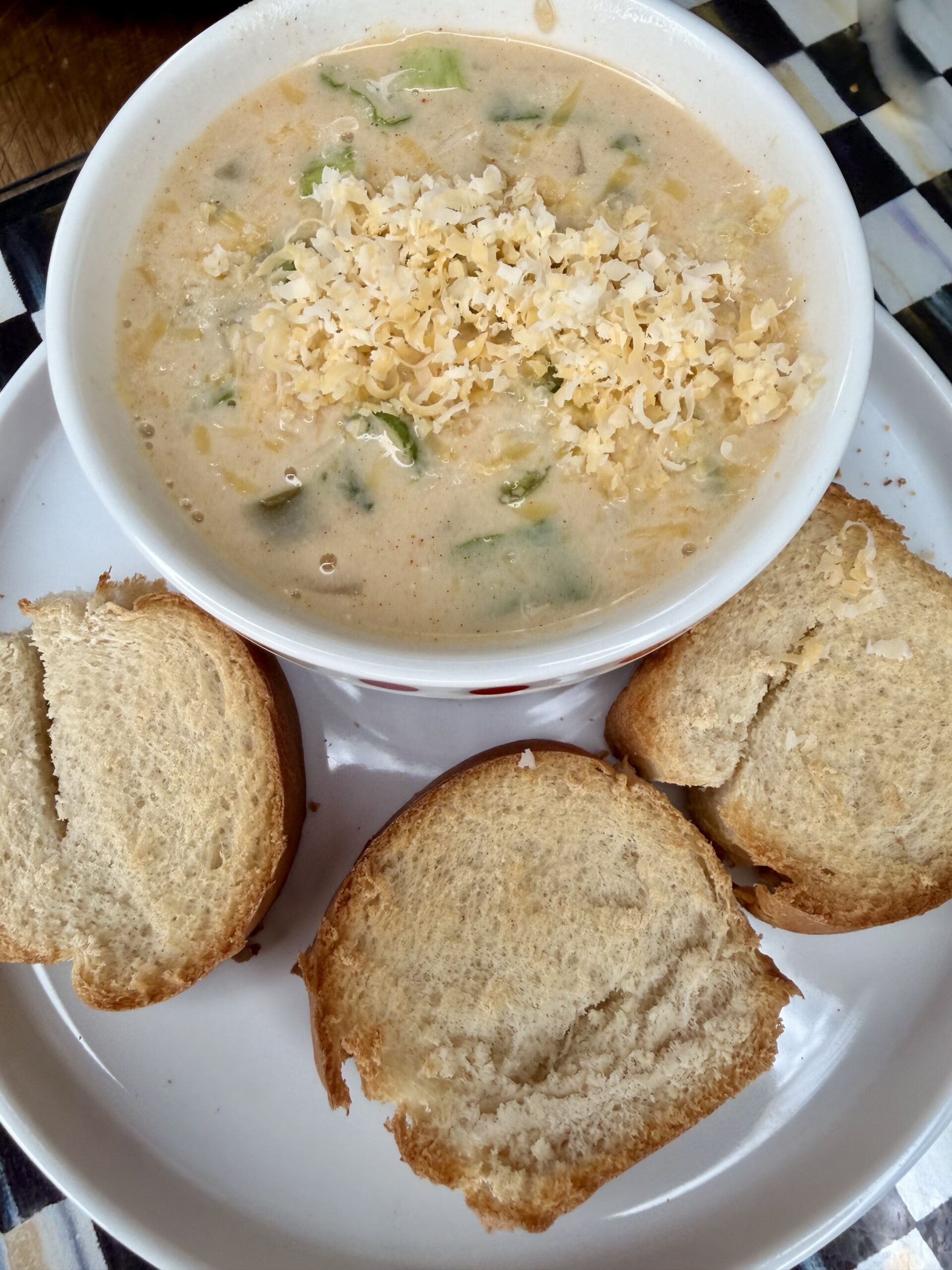 A bowl of creamy soup topped with shredded cheese, served with three pieces of sliced bread on a white plate. The soup contains visible green vegetable pieces.
