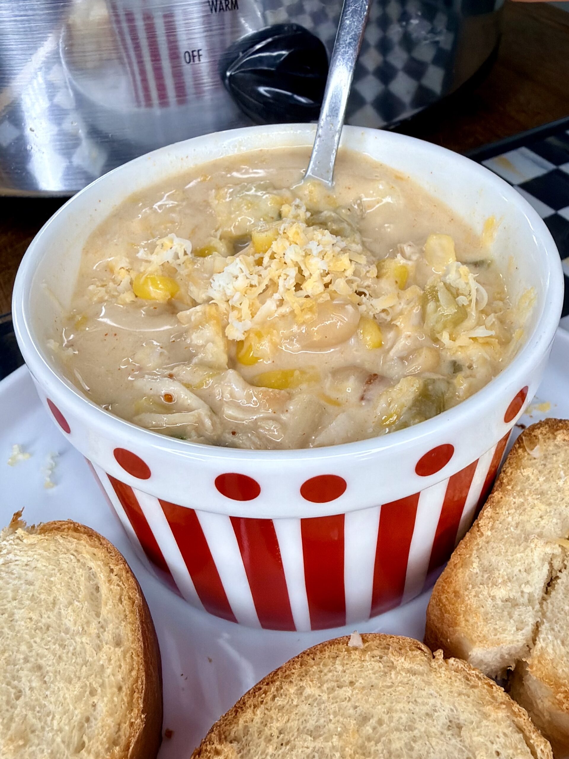A bowl of creamy soup with beans, corn, and cheese sits in a red-and-white striped bowl, garnished with shredded cheese, and served with slices of toasted bread. A slow cooker is visible in the background.