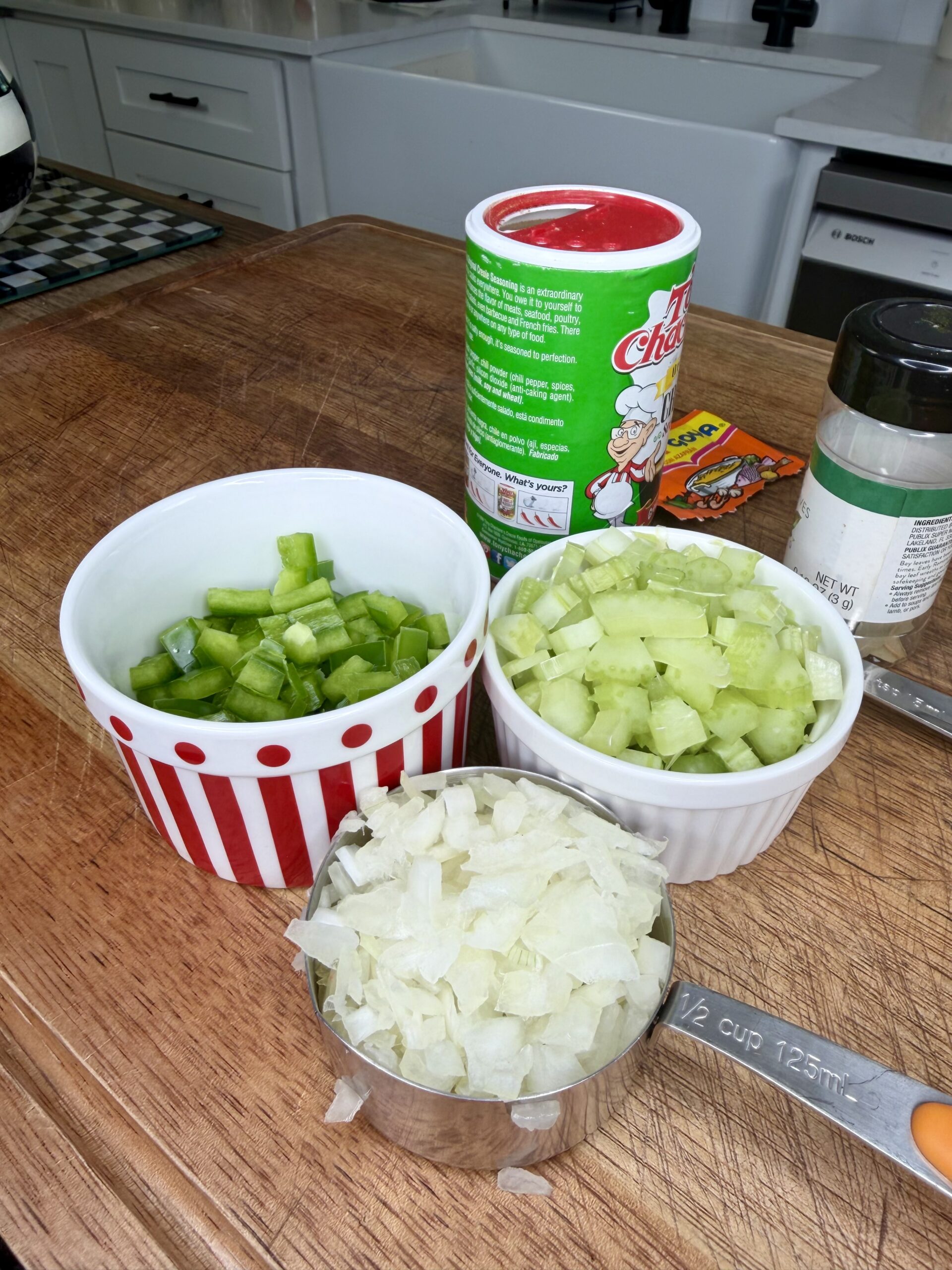 Diced green bell pepper, celery, and onion are in bowls and a measuring cup on a wooden cutting board. Seasoning containers, including Tony Chachere’s, are nearby in a bright kitchen.
